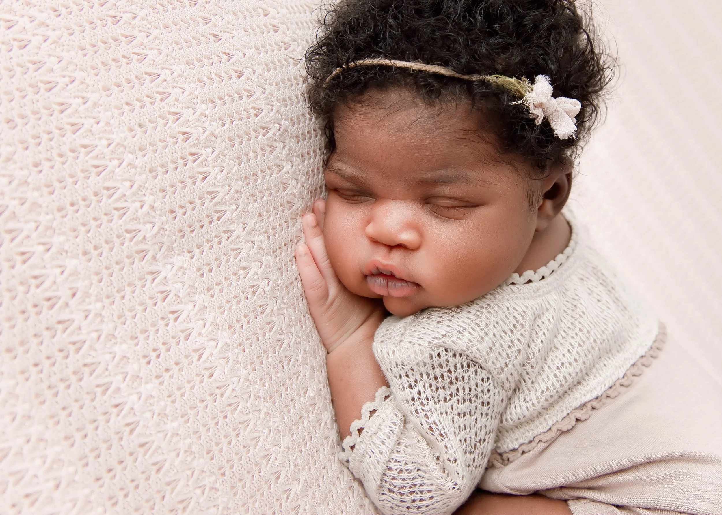 A sleeping baby girl with dark curly hair and a cream-colored headband, resting her cheek on a textured, creamy-colored blanket.