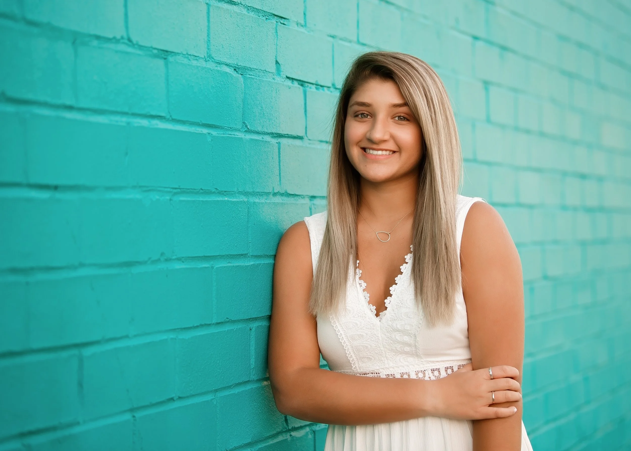 A young woman with long blonde hair smiling and standing against a turquoise brick wall, wearing a white sleeveless dress and a silver necklace.