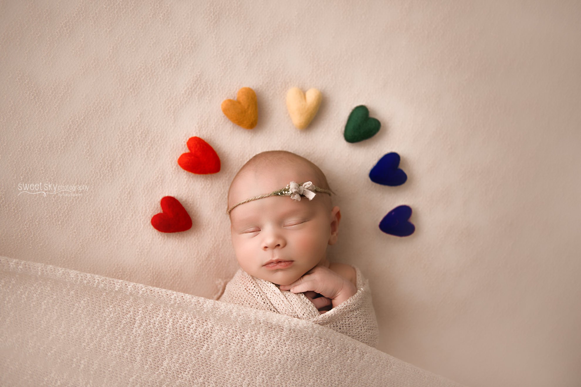A sleeping baby wrapped in a beige blanket, wearing a headband, surrounded by rainbow-colored heart-shaped felt decorations.