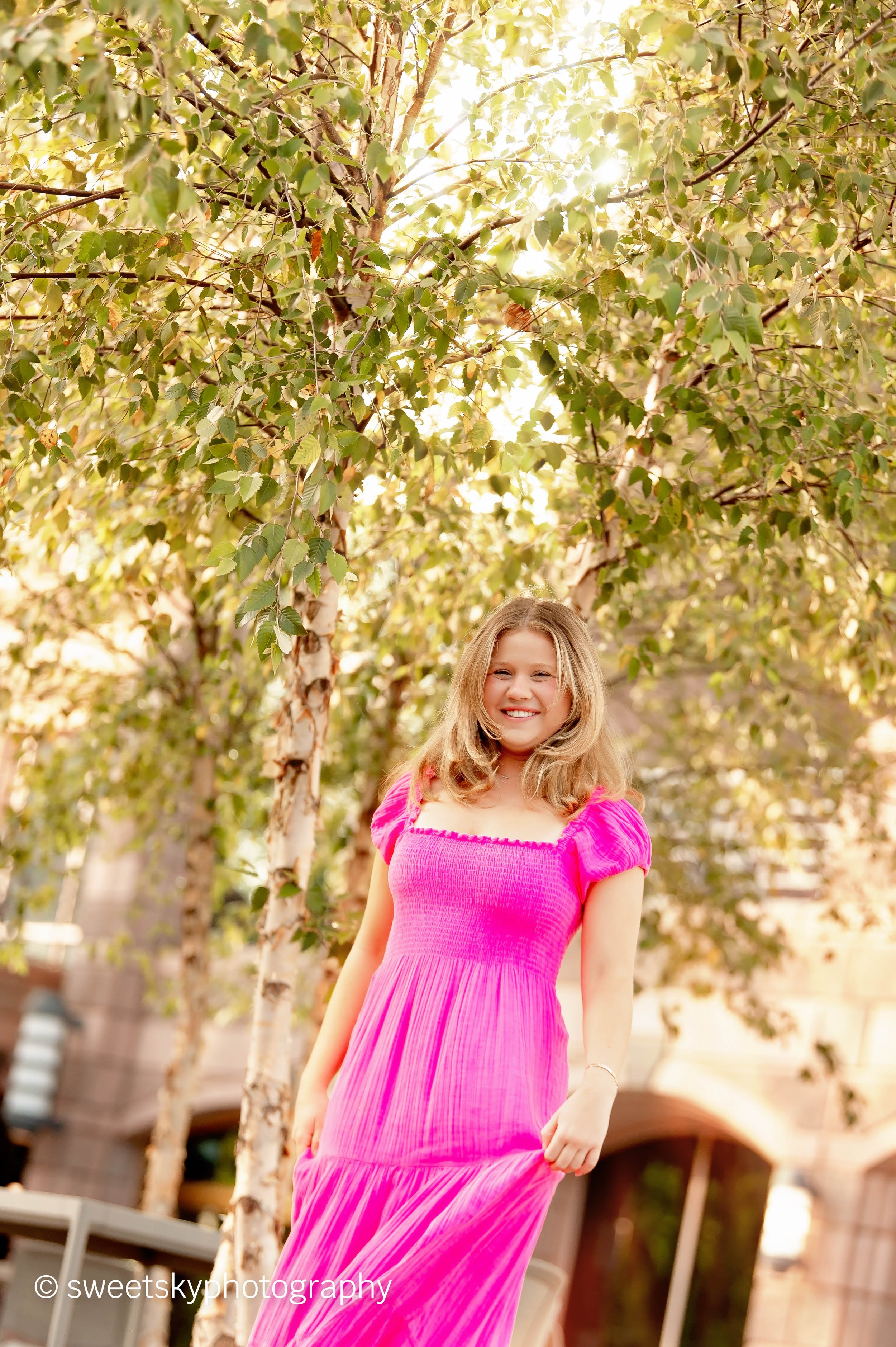 A woman with blonde hair wearing a bright pink dress, smiling and standing outdoors under a tree with green leaves and sunlight shining through.