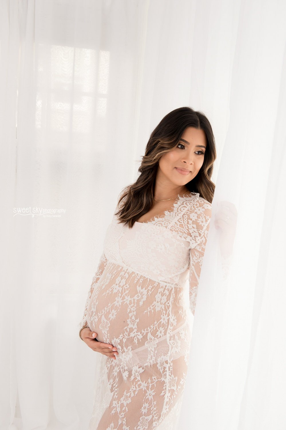 A pregnant woman in a sheer lace white dress gently holding her belly, standing by sheer white curtains, looking thoughtfully to the side.