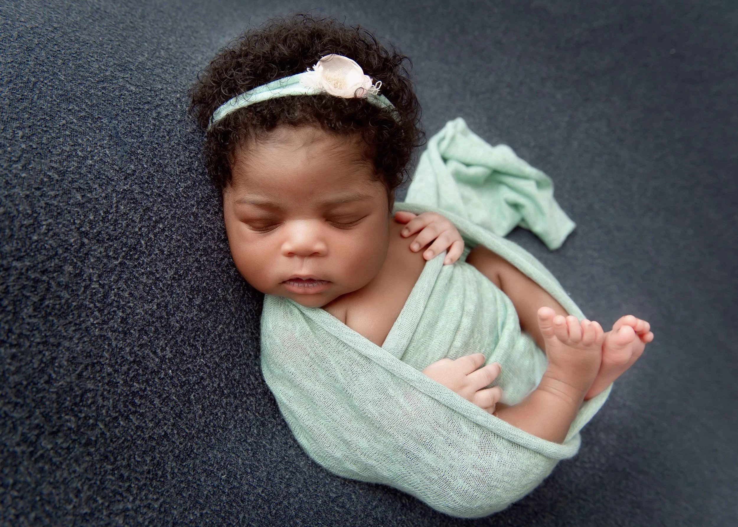 A sleeping baby girl with curly hair, wrapped in a light green blanket, laying on a dark textured surface, wearing a white headband with a flower detail.
