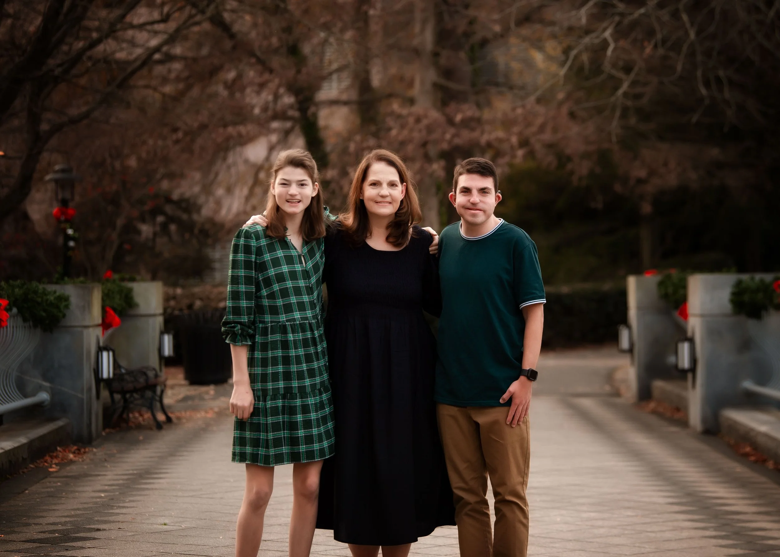 A woman with dark brown hair standing between a teenage girl with brown hair in a green plaid dress and a teenage boy with short dark hair in a dark green shirt, all smiling outdoors in a park with autumn trees and red decorations.