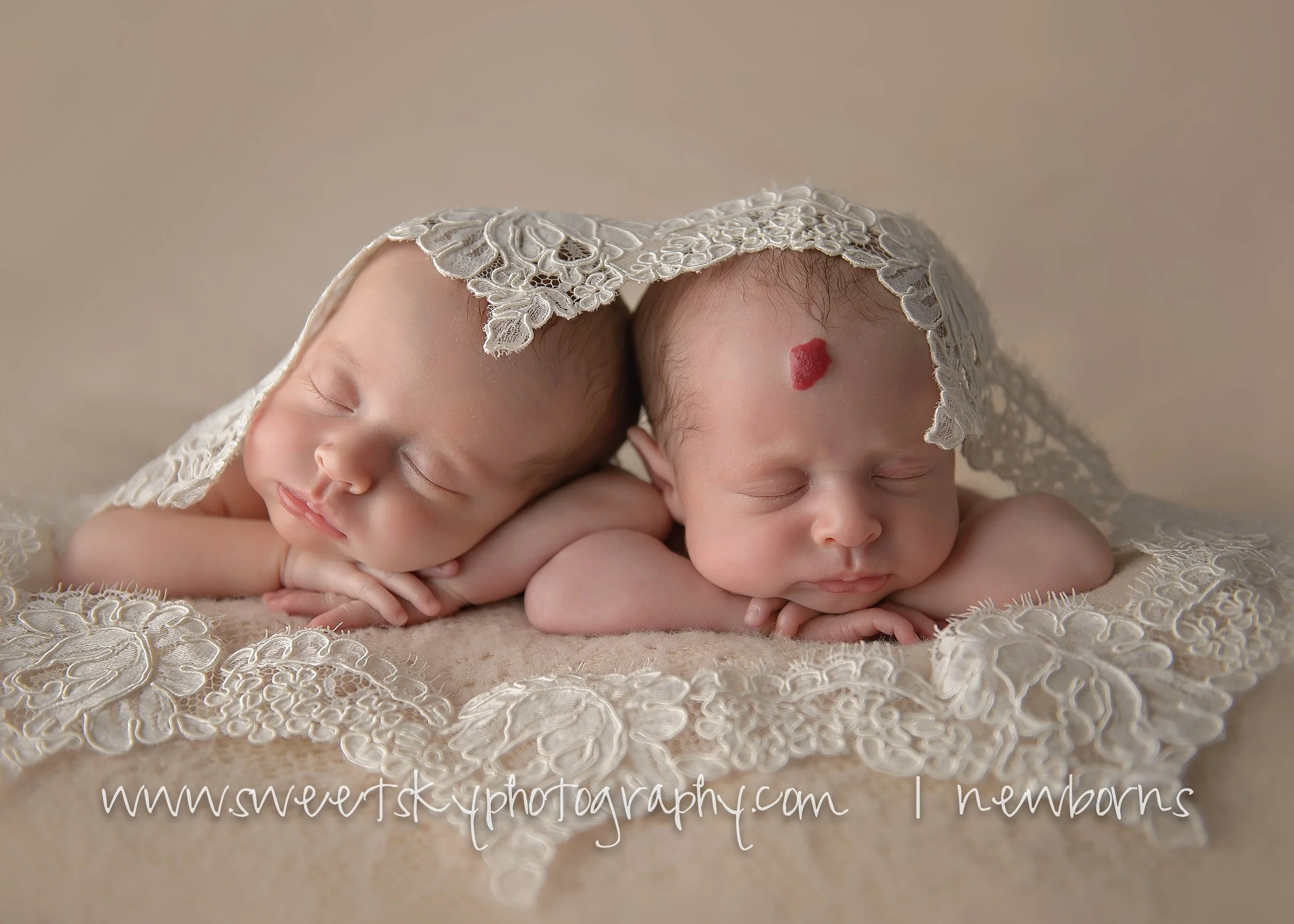 Two newborn babies sleeping with lace cloth draped over their heads, resting on a soft beige surface.