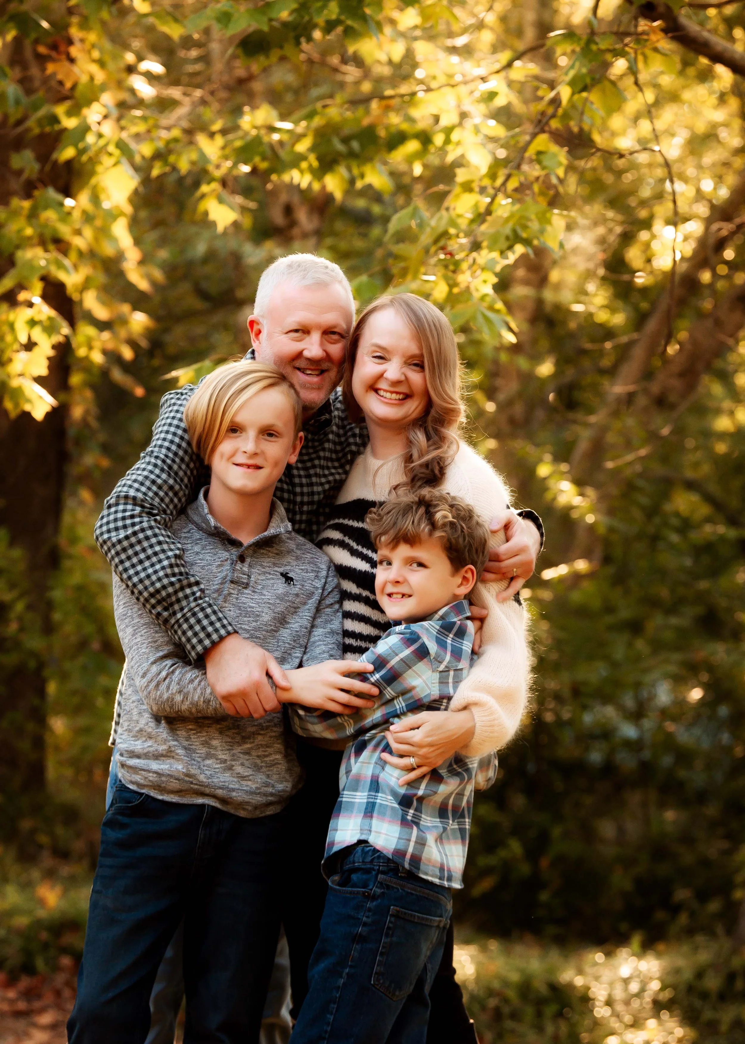 A family of five hugging outdoors in a wooded area during fall, smiling and enjoying the moment.