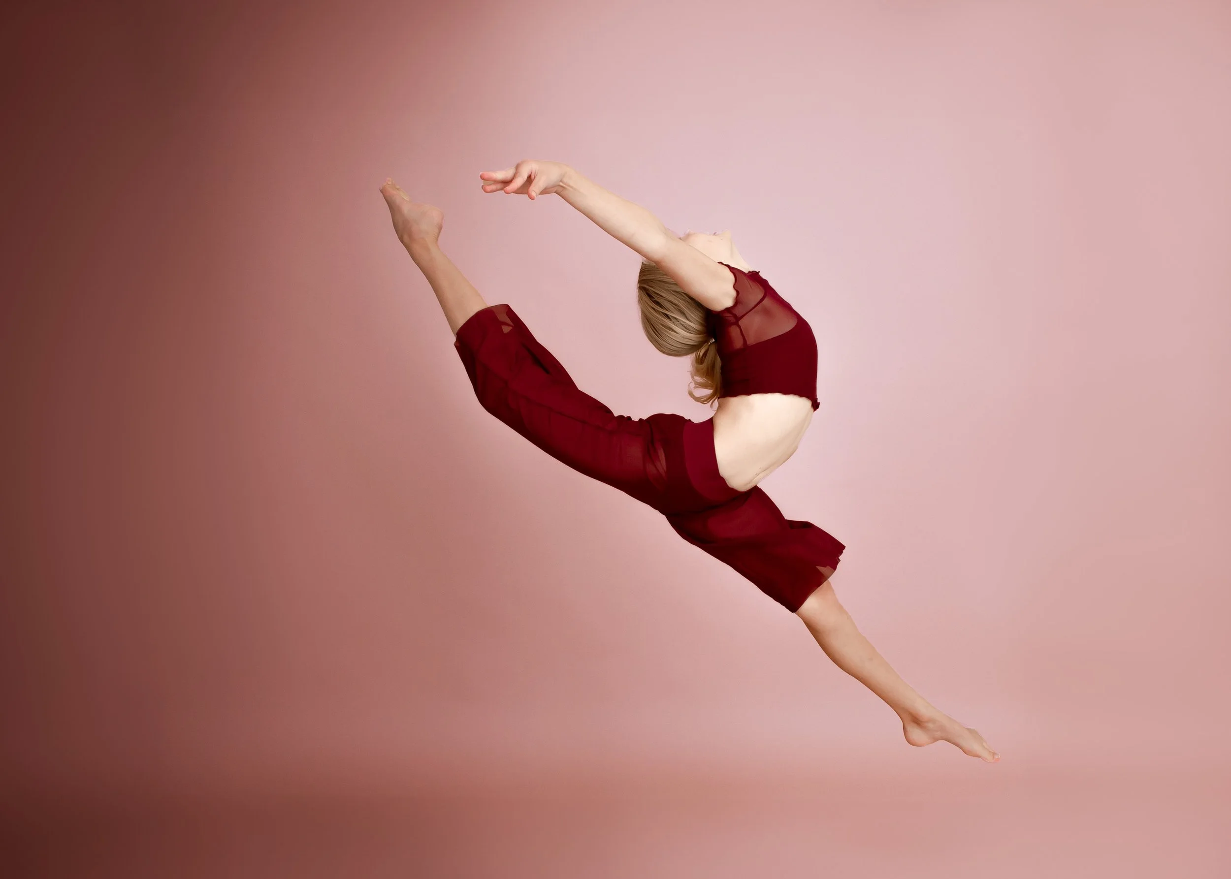 A woman in red clothing performing a yoga pose with her body stretched diagonally, arms extended overhead, and in front of a pink background.
