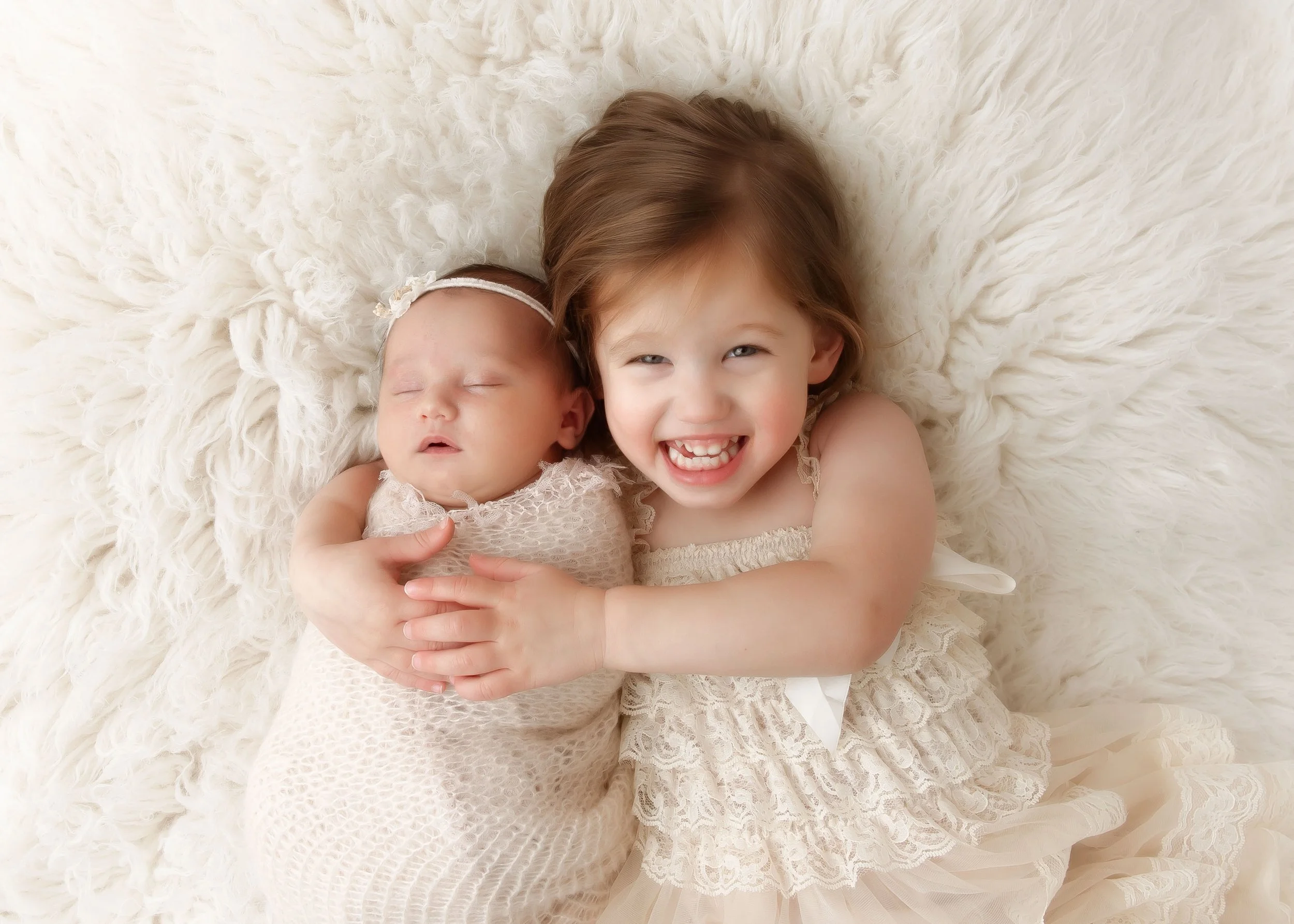 A young girl with brown hair and a wide smile holding a sleeping baby girl on a fluffy white surface.