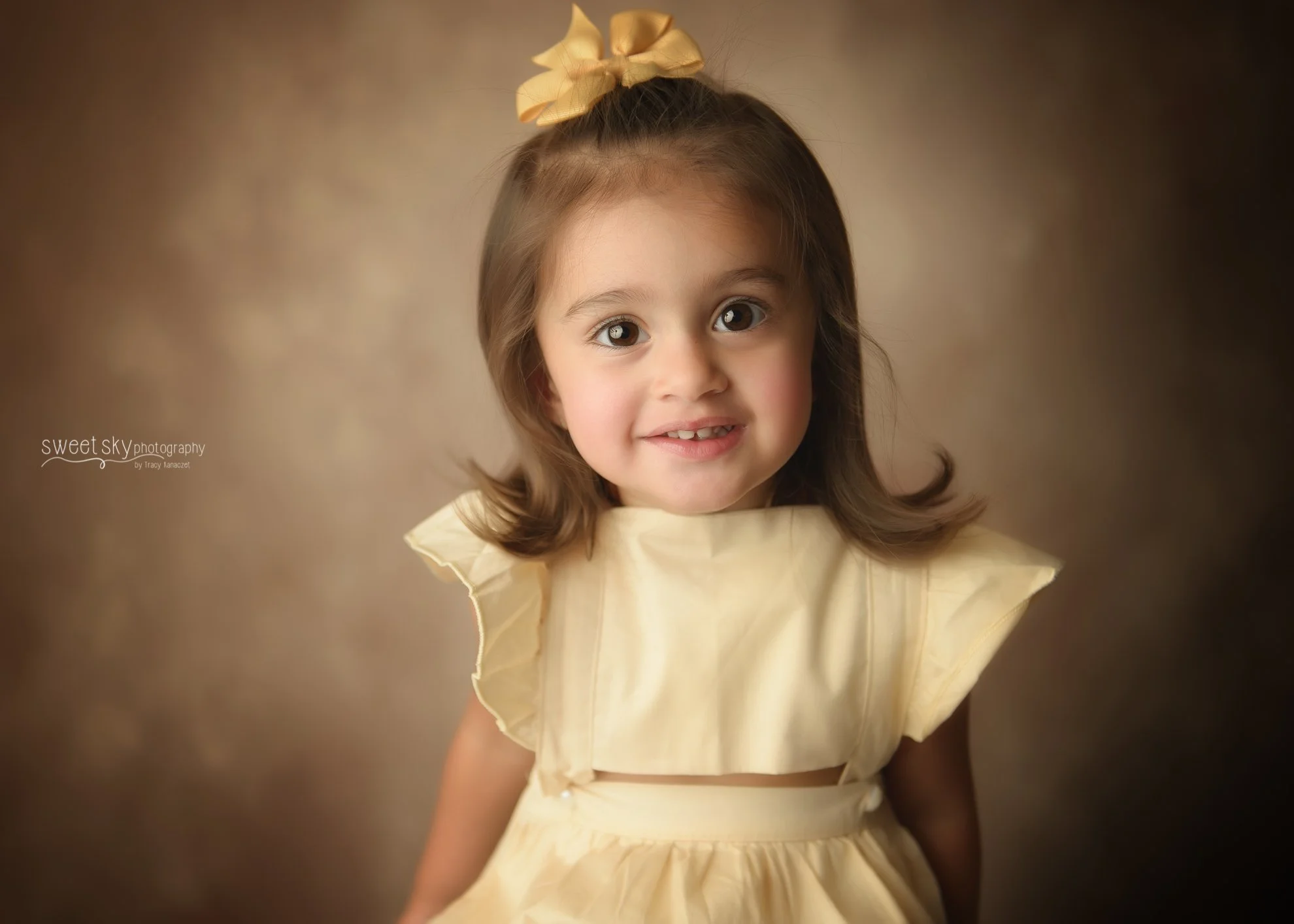 A young girl with brown hair, big brown eyes, and a big smile, wearing a yellow dress and a matching yellow bow in her hair, standing against a brown background.