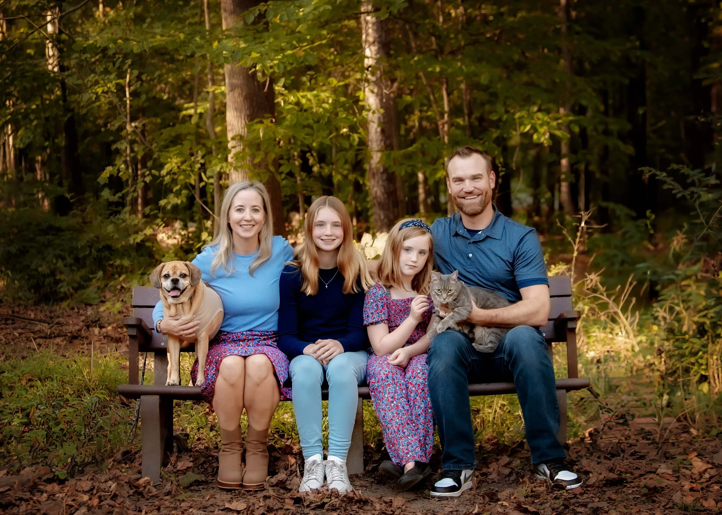 A family of five and their two pets sit on a park bench in a wooded area, smiling at the camera.