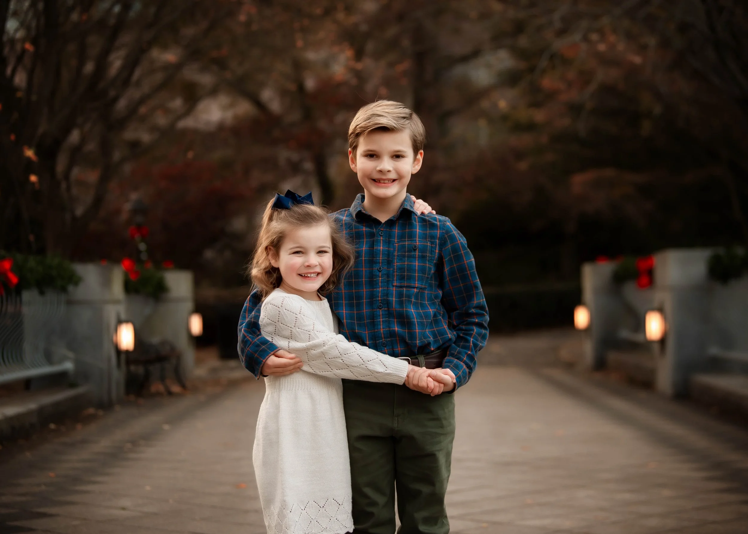 A young boy and girl smiling and holding hands on a bridge with festive holiday lights and decorations, during autumn.