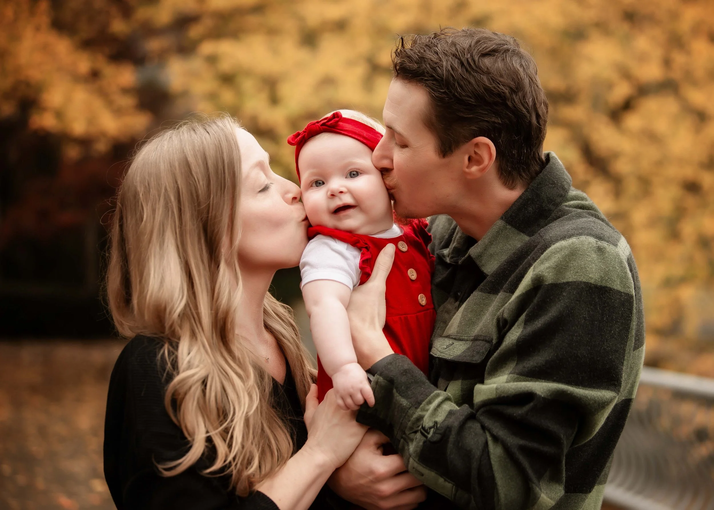 A family of three, including a mother, father, and their baby girl, sharing kissing moments outdoors during fall, with colorful autumn leaves in the background.