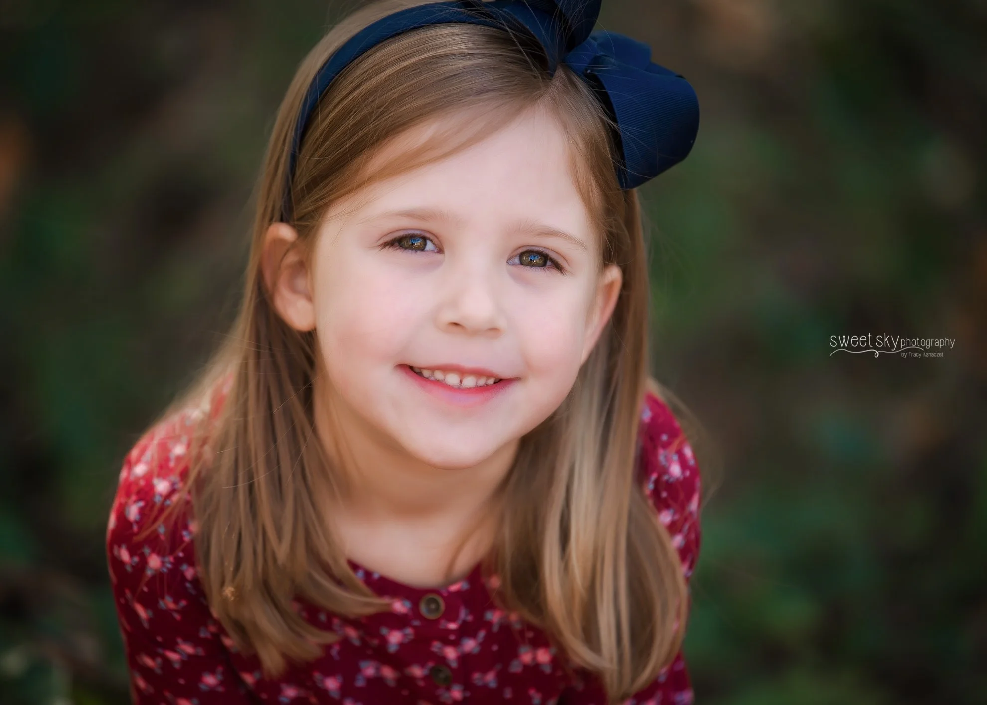 Close-up of a young girl with long light brown hair, wearing a blue headband and a red floral dress, smiling outdoors with a blurred green background.