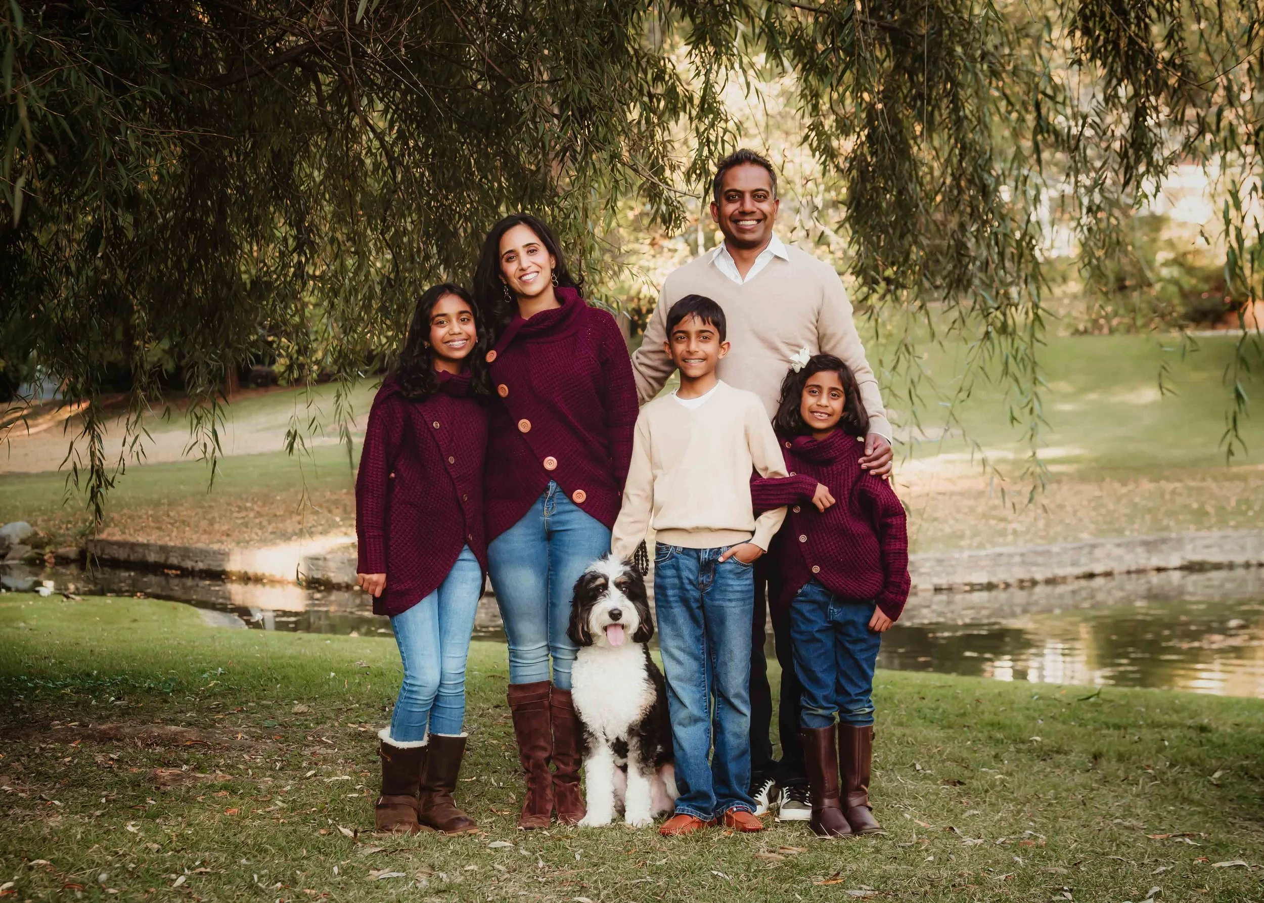 Family of six outdoor by a pond in autumn, smiling for a photo. Two adults, three children, and a dog.