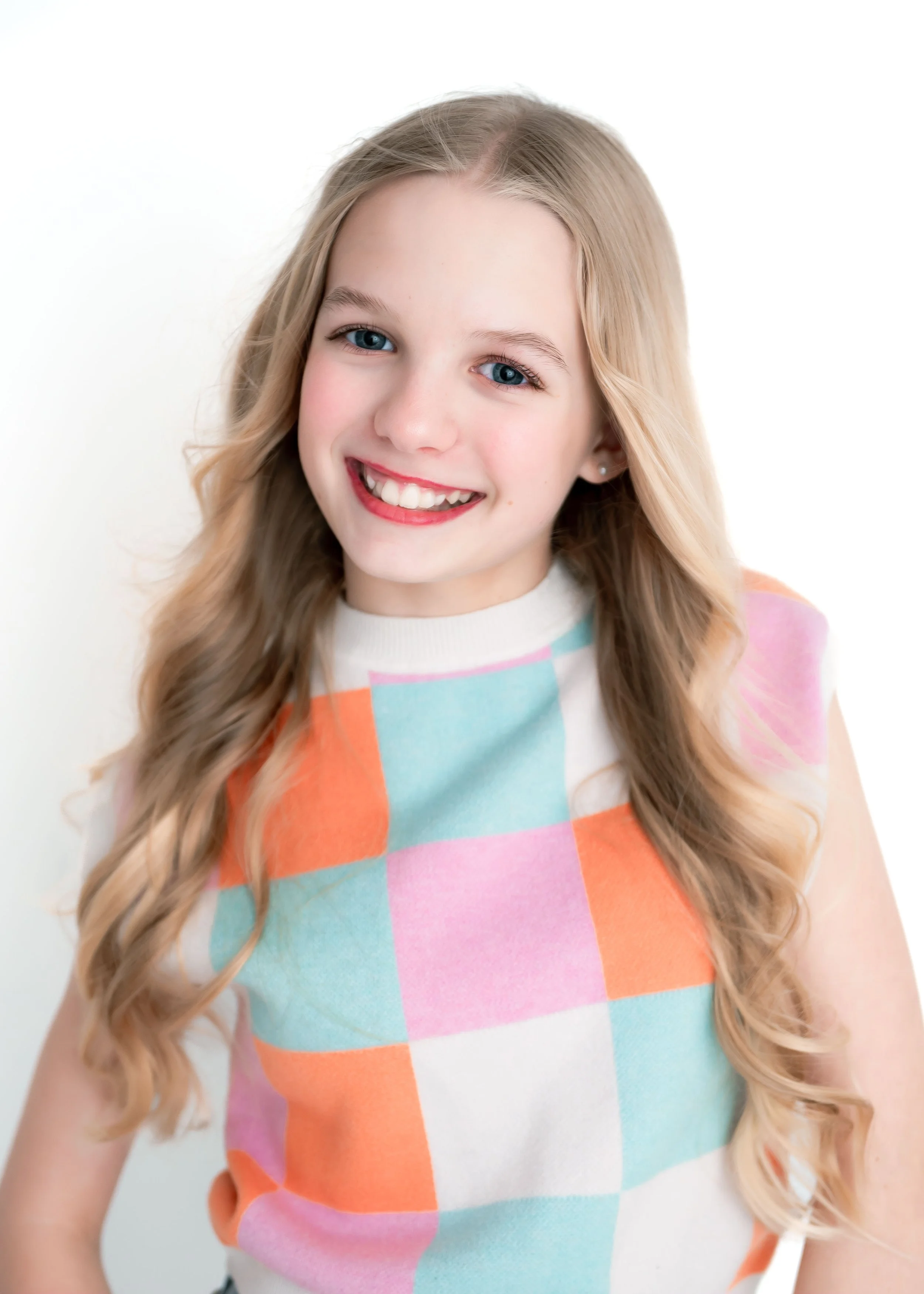 A young girl with long, wavy blonde hair and blue eyes, smiling, wearing a sleeveless, pastel-colored checkered top, standing against a white background.