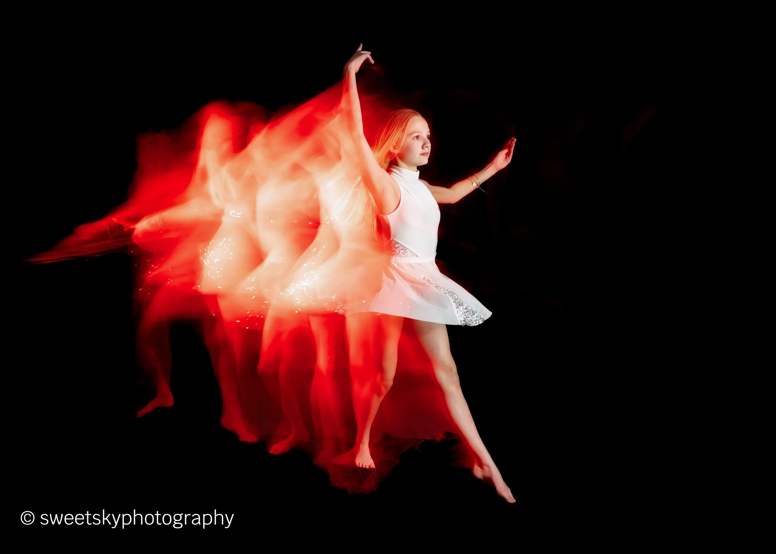 A young girl in a white dress performing a dance move against a black background, with a motion blur effect creating multiple positions of her arms, legs, and dress.