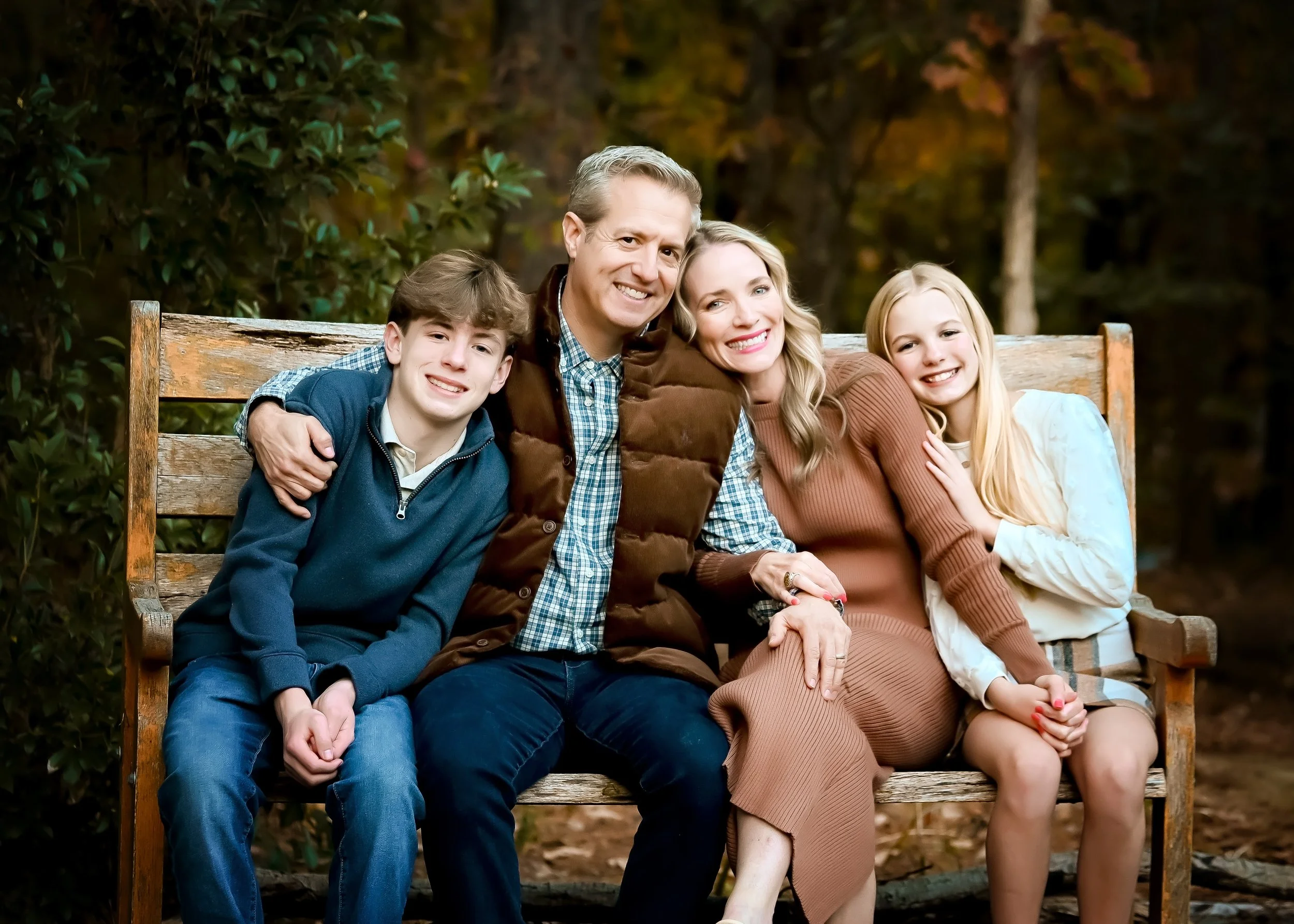Family sitting on a wooden bench outdoors during fall, smiling at the camera.