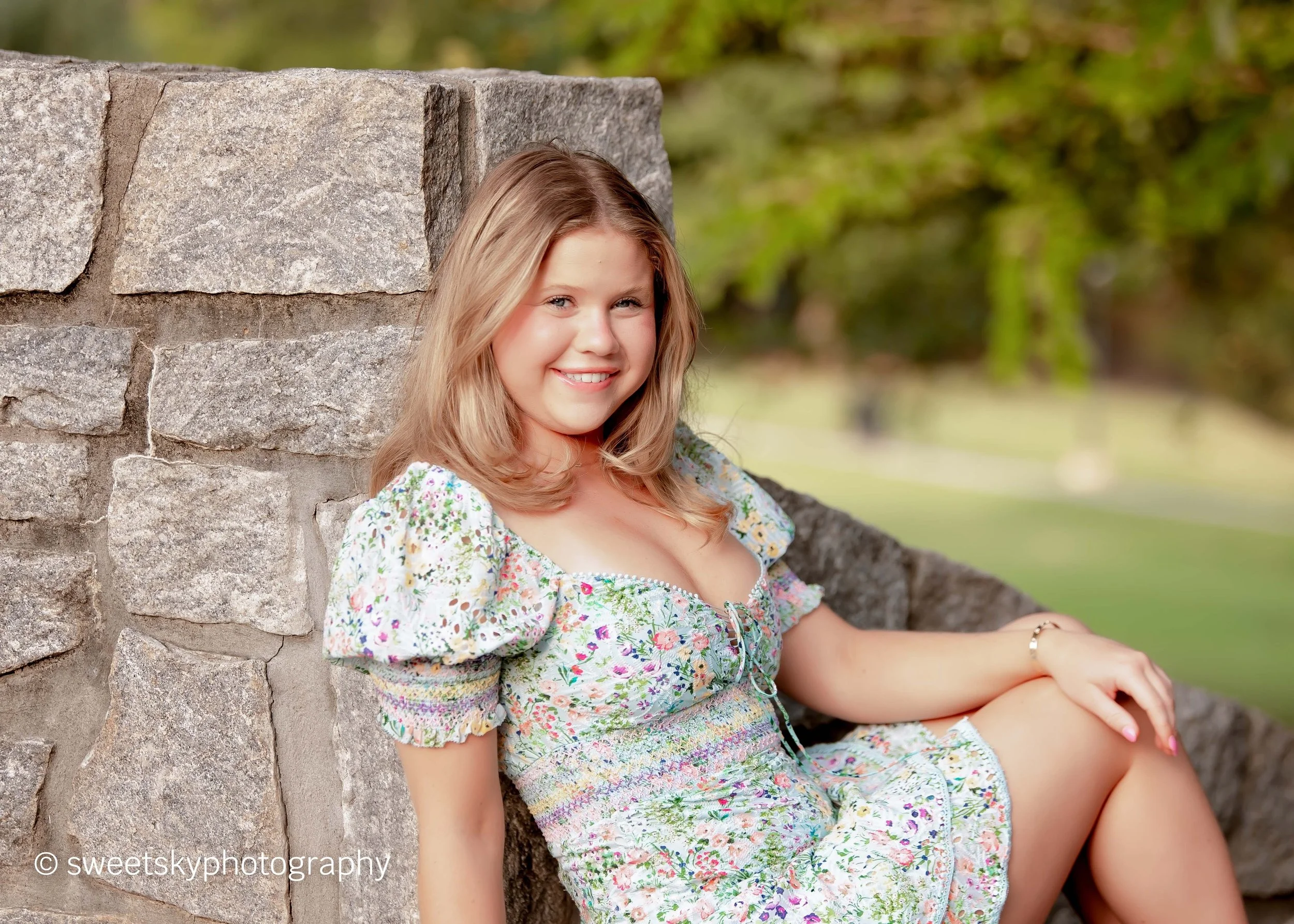 A young woman with blonde hair, wearing a floral dress with puffed sleeves, sitting on a stone ledge outdoors, smiling at the camera, with green trees and grass in the background.