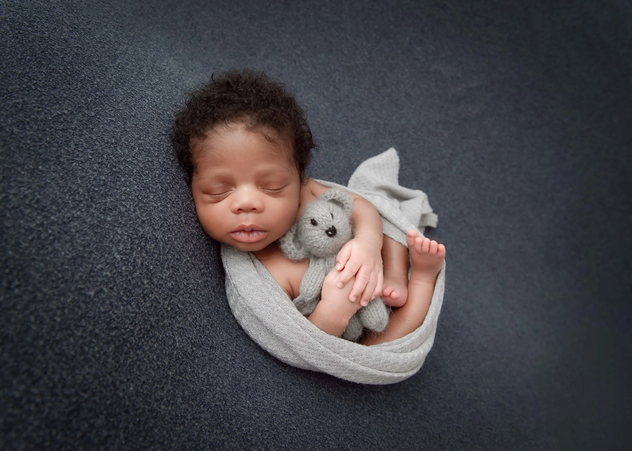 A sleeping newborn baby with curly hair, cuddling a small, gray, stuffed bear on a dark textured surface.