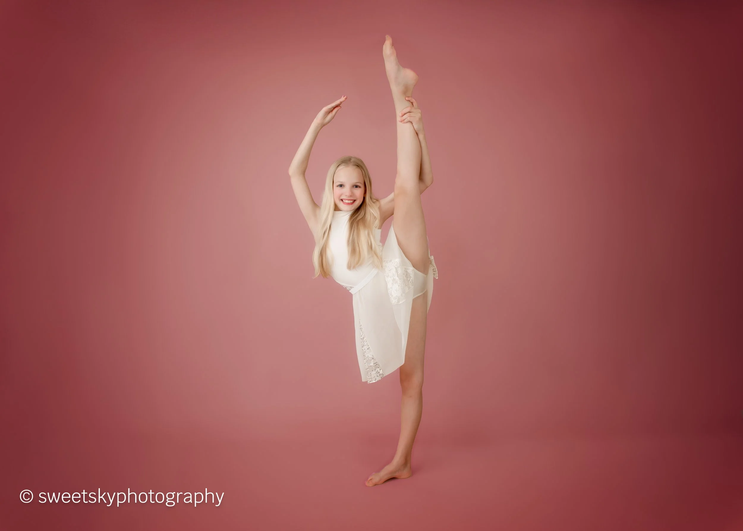 Young girl in a white dress performing a yoga pose with one leg raised and a big smile, standing on a pink background.