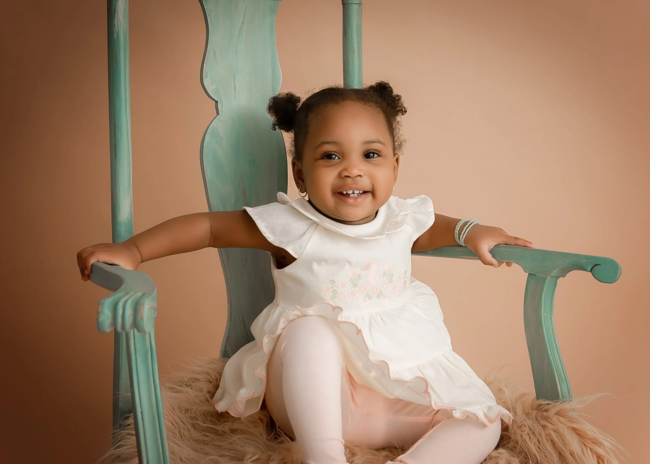 A smiling young girl with two pigtails sitting on a pastel blue wooden high-back chair, wearing a white dress with pink floral embroidery, white tights, and bracelets, on a soft, furry blanket against a neutral background.