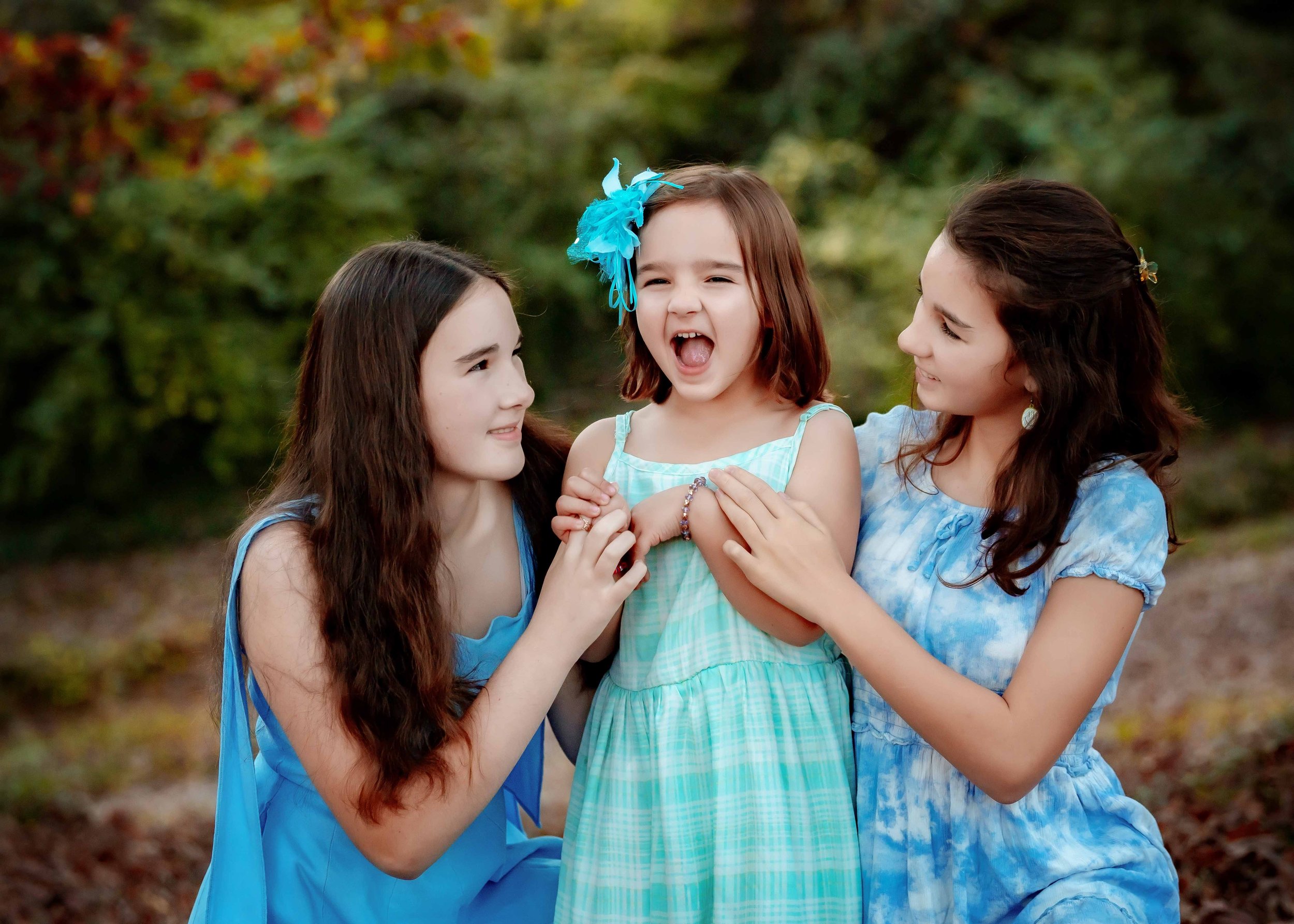 Two young women holding and smiling at a cheerful girl in a green dress with a blue flower hair accessory, outdoors with trees in the background.
