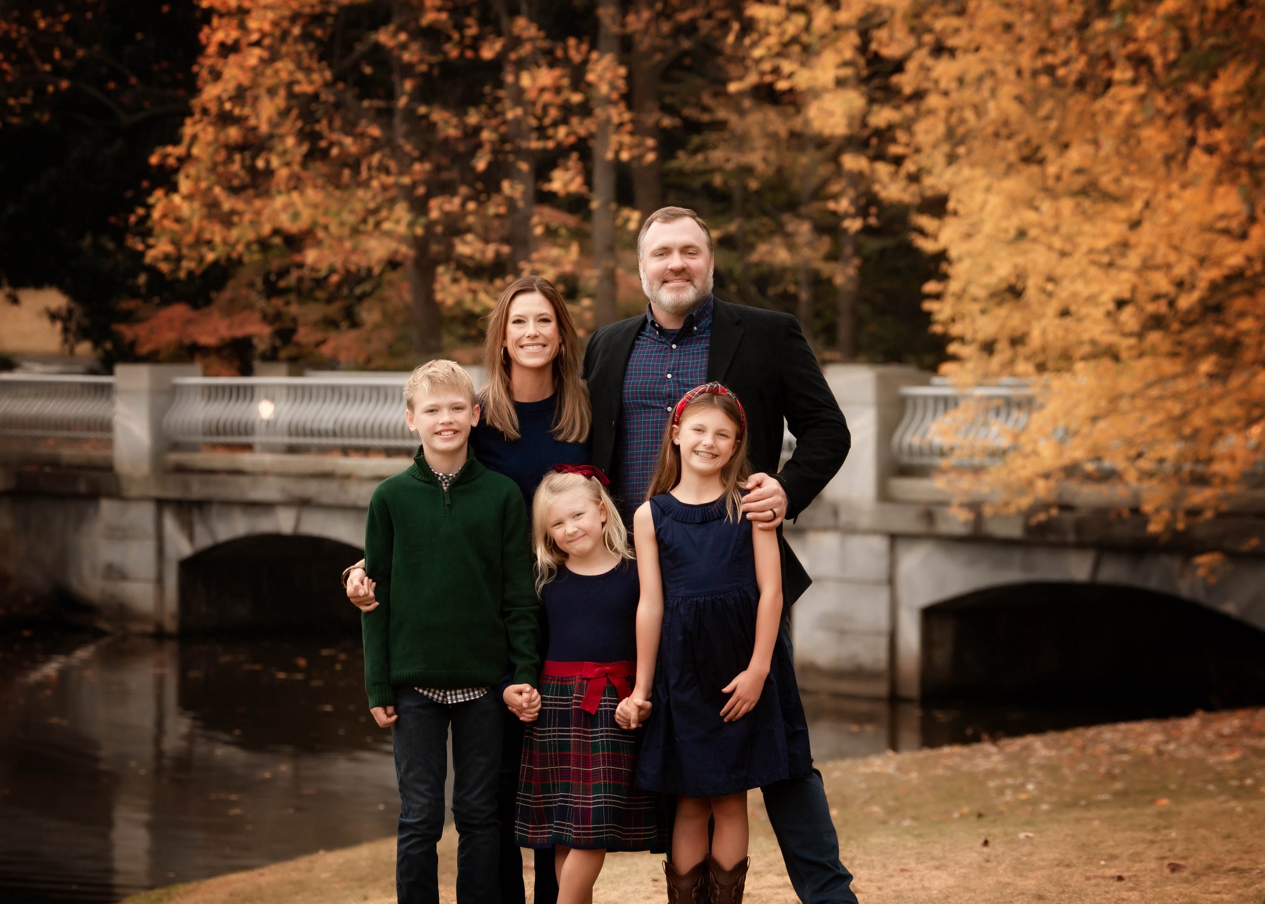 A family of six posing outdoors near a stone bridge with autumn trees in the background.