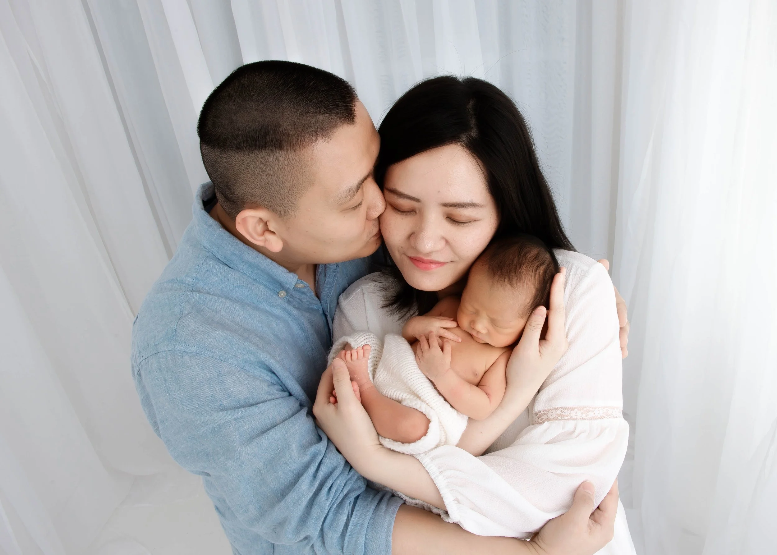 A couple holding their newborn baby, with the father giving a gentle kiss on the mother's forehead.