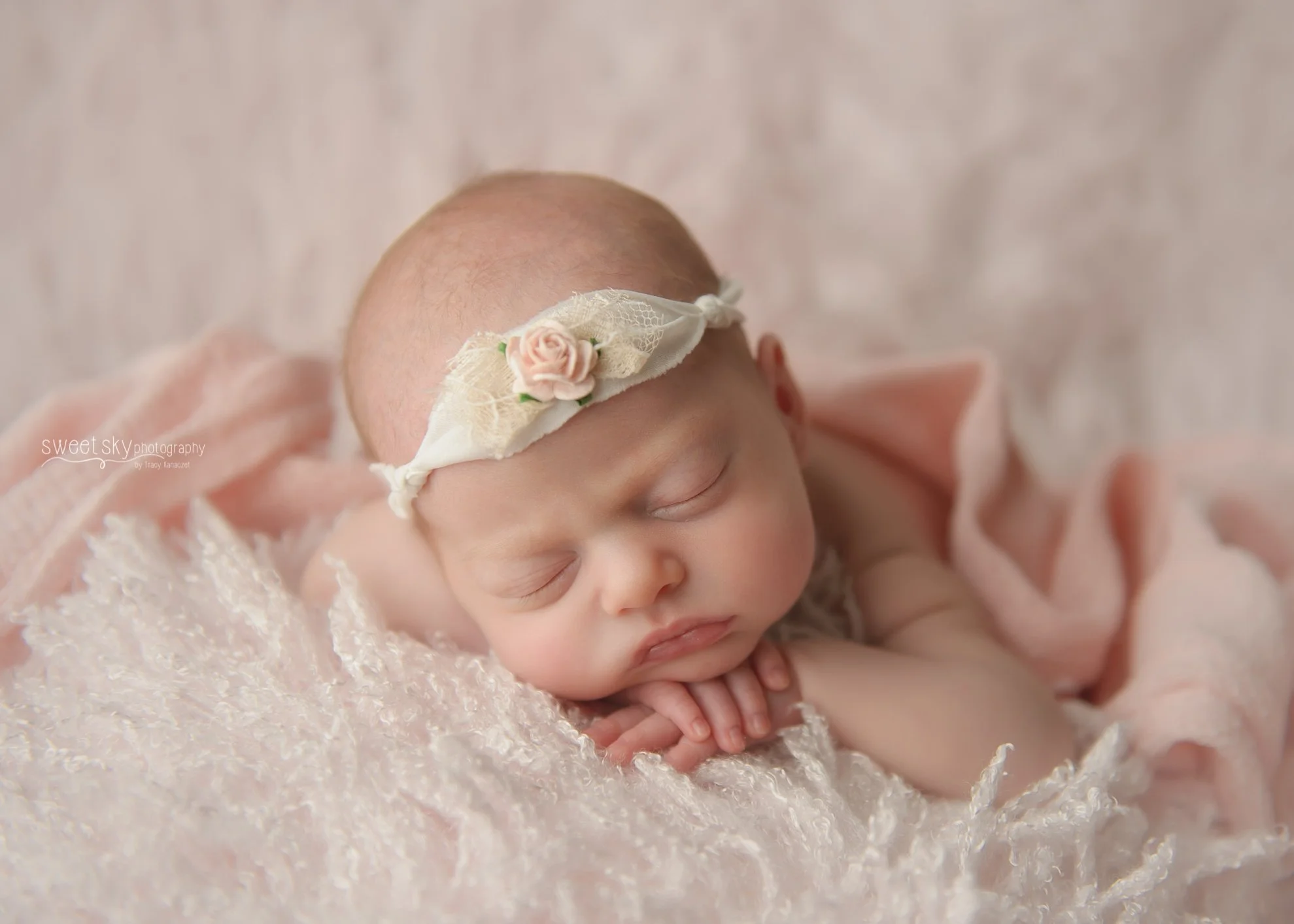 A sleeping newborn baby with a pink flower headband, lying on a soft, textured pink blanket with hands tucked under chin.