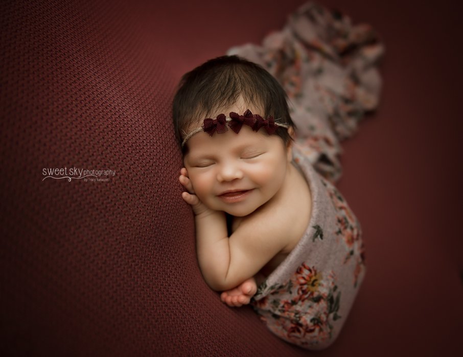 Close-up of a smiling baby girl with dark hair, wearing a floral headband and swaddled in a floral blanket, lying on a maroon textured surface.