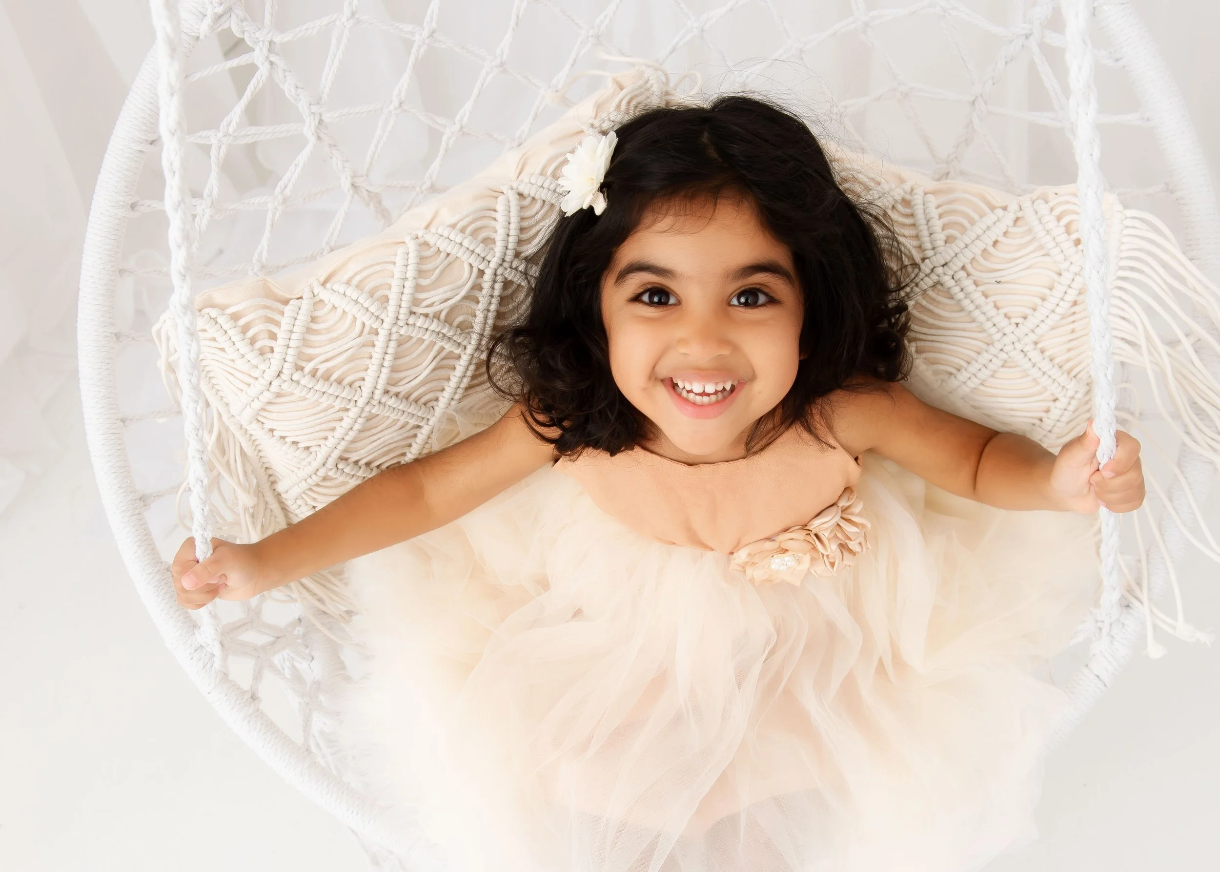 A young girl with dark curly hair, smiling and looking up, sitting in a white hanging chair wearing a peach-colored dress with tulle. She has a white flower in her hair.