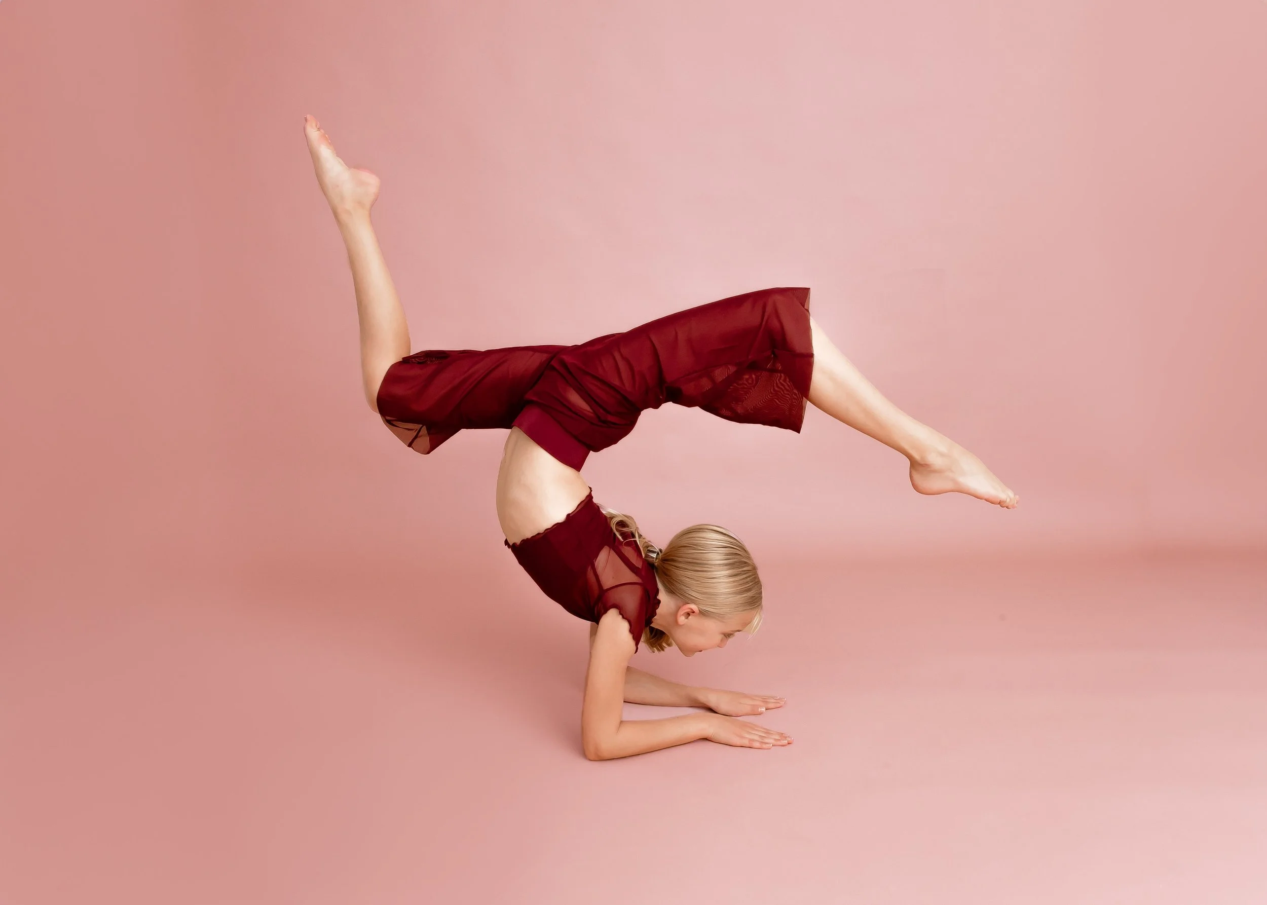 A woman in maroon clothing performing a yoga pose on a pink background, balancing on her forearms with her legs extended over her head.