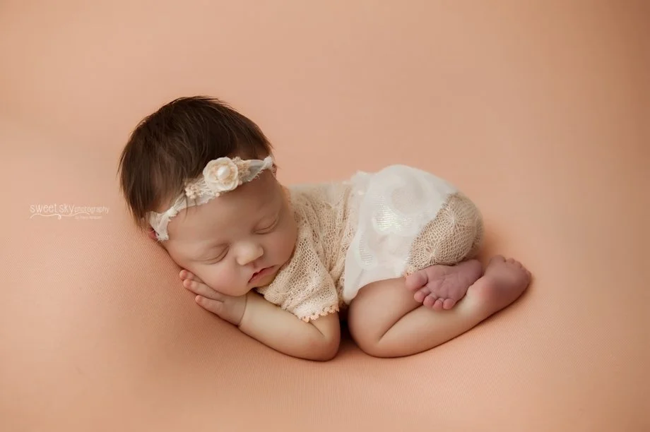 A sleeping baby girl with a floral headband, wearing a lace dress and a white skirt, curled up in a fetal position on a peach-colored background.