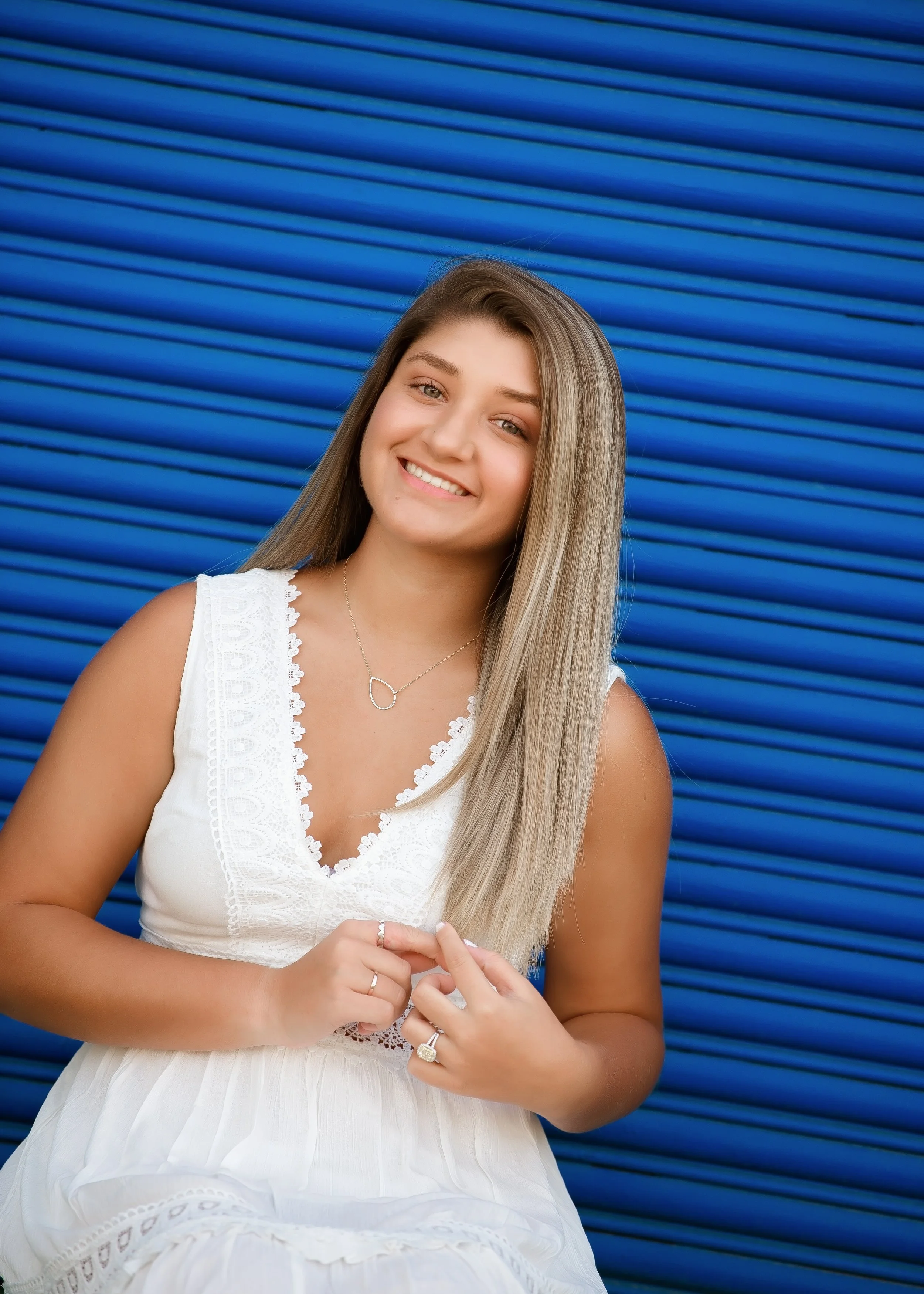 A young woman with long blonde hair smiling, wearing a white sleeveless dress with lace details, standing in front of a blue corrugated metal wall.