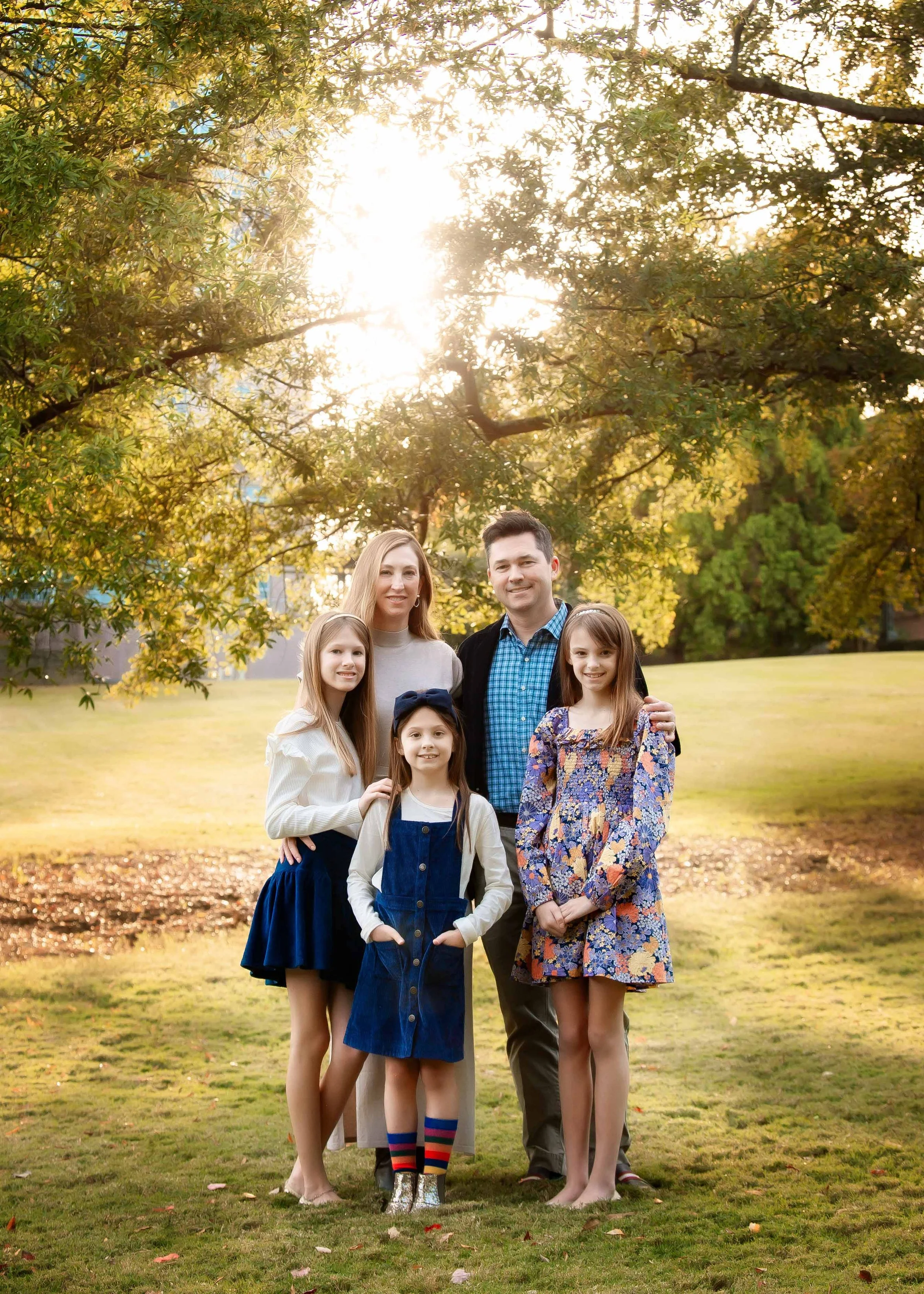 A family of five standing outdoors in a park during late afternoon or early evening, with sunlight filtering through trees in the background. The family includes a woman, a man, and three young girls, all smiling and dressed in casual clothes.