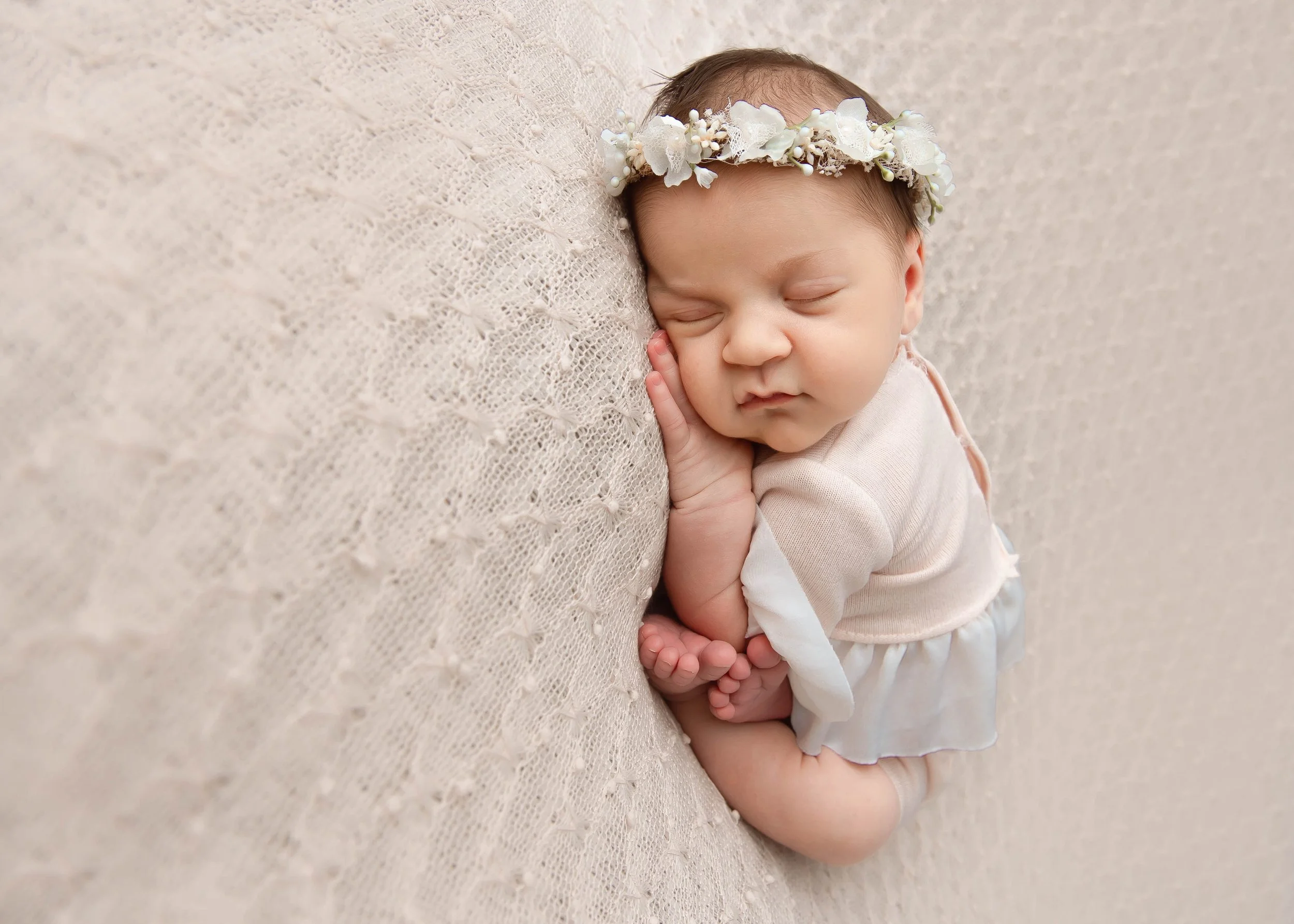 A sleeping baby girl with a floral headband, laying on her side on a textured beige blanket.