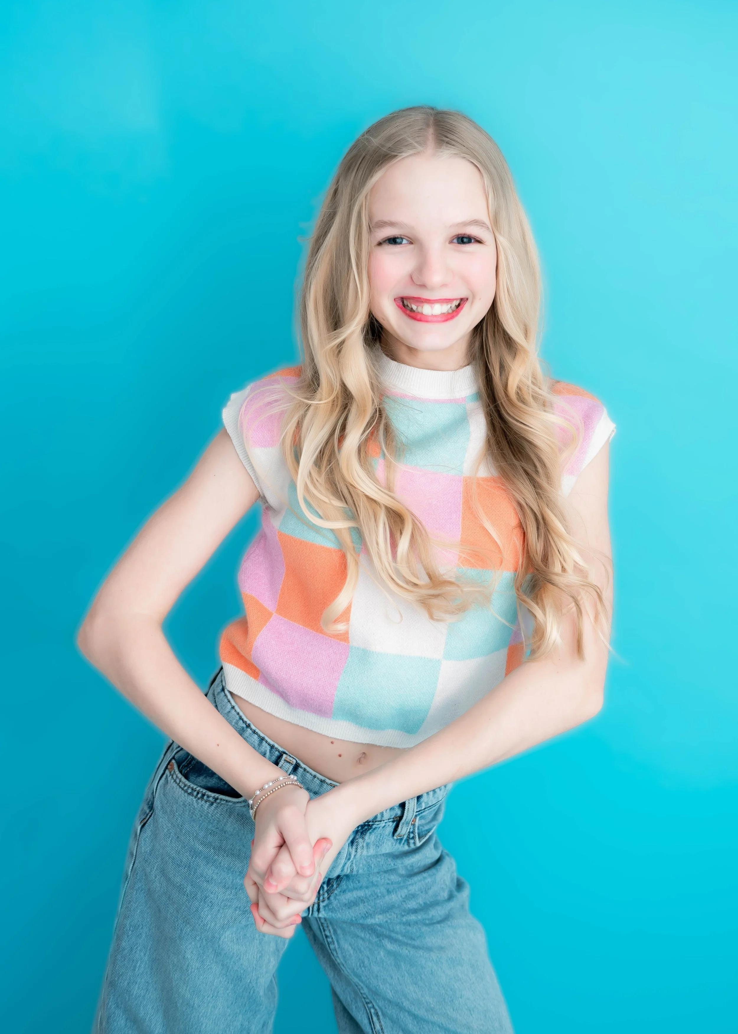 A young girl with long blonde hair, smiling, wearing a colorful pastel checkered sleeveless top and blue jeans, standing against a bright blue background.