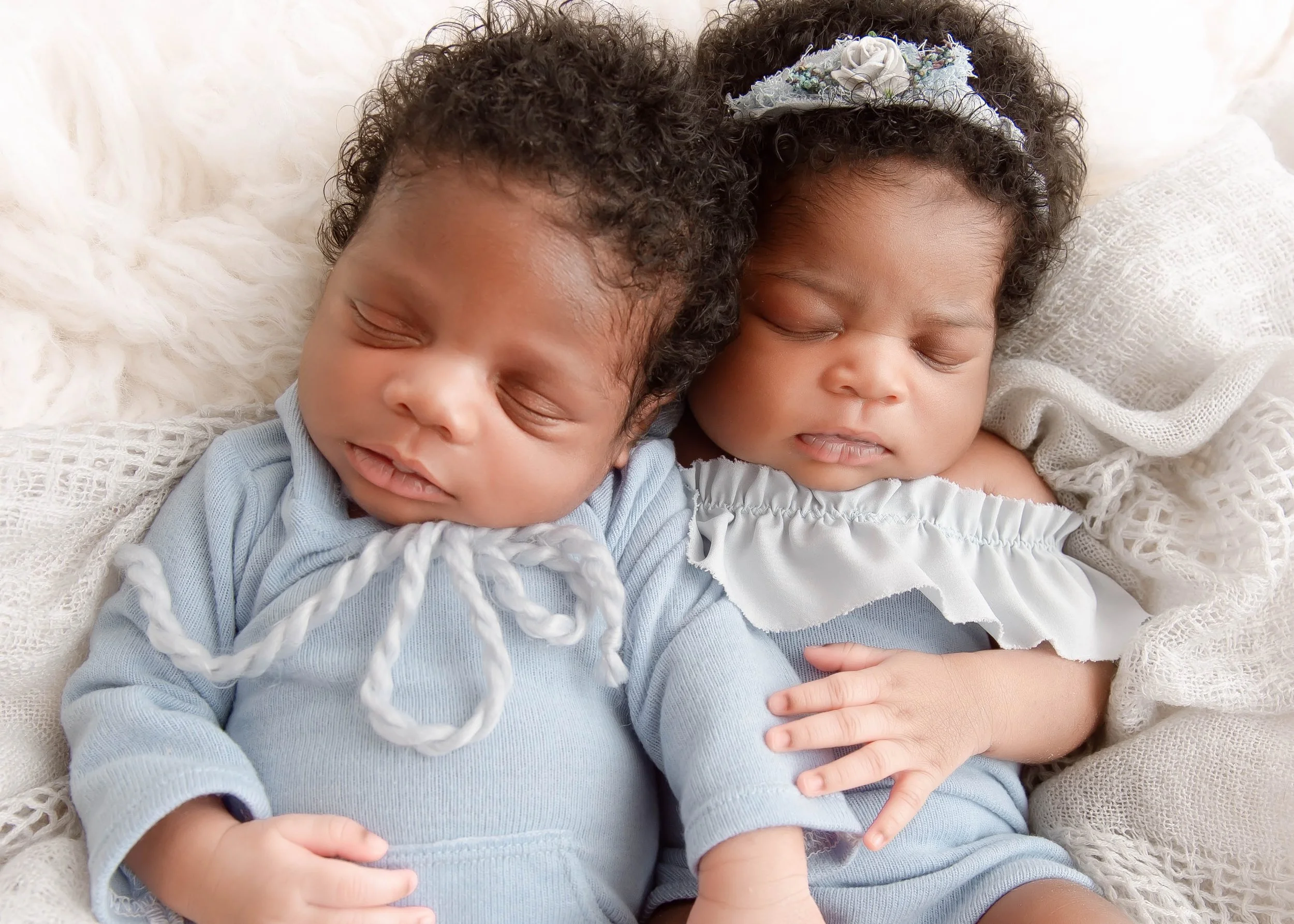 Two sleeping babies with curly hair, one wearing a blue outfit and the other a light-colored dress, lying close together on a soft, textured white blanket.