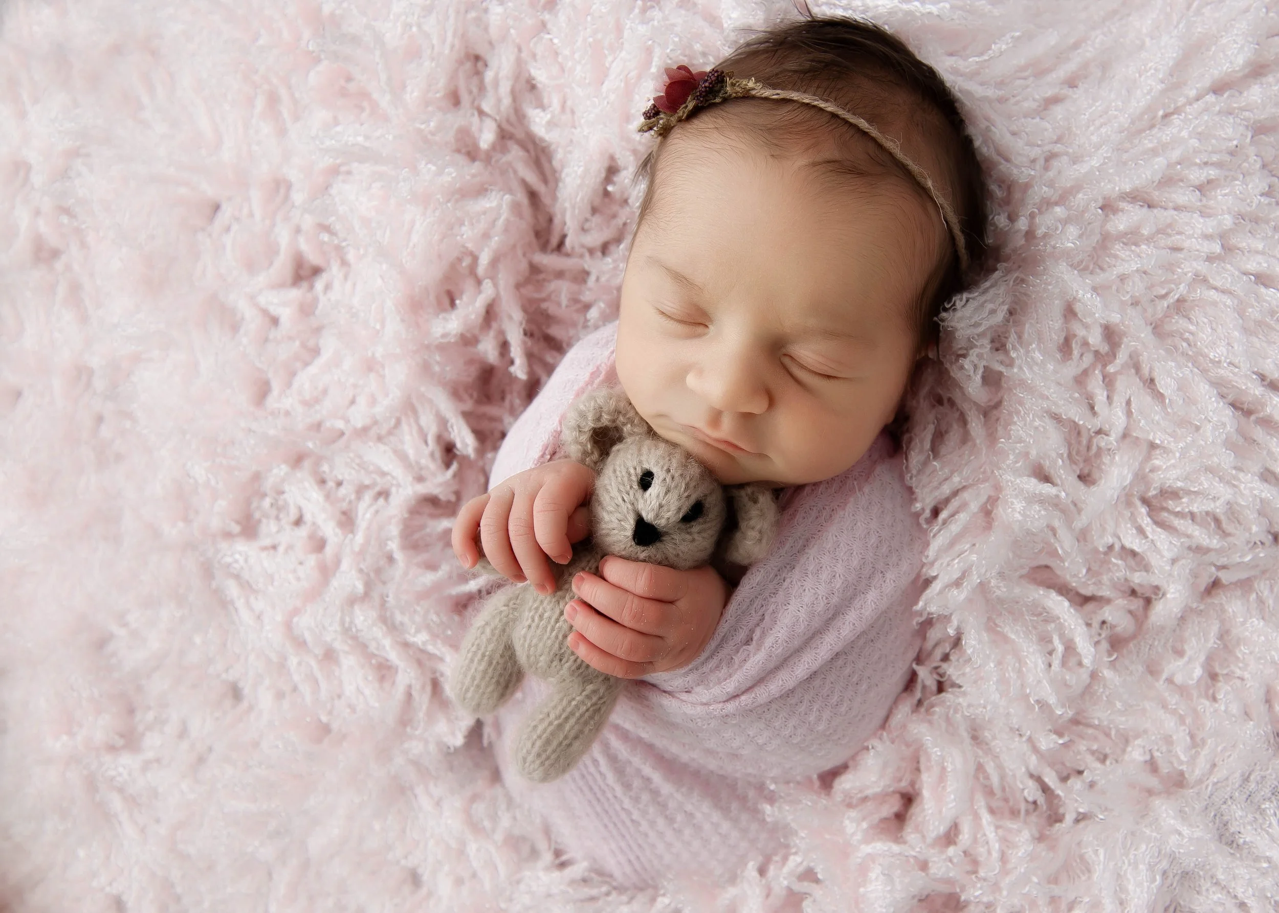 A sleeping baby girl with a pink headband, cuddling a small knit gray teddy bear on a soft pink fluffy blanket.
