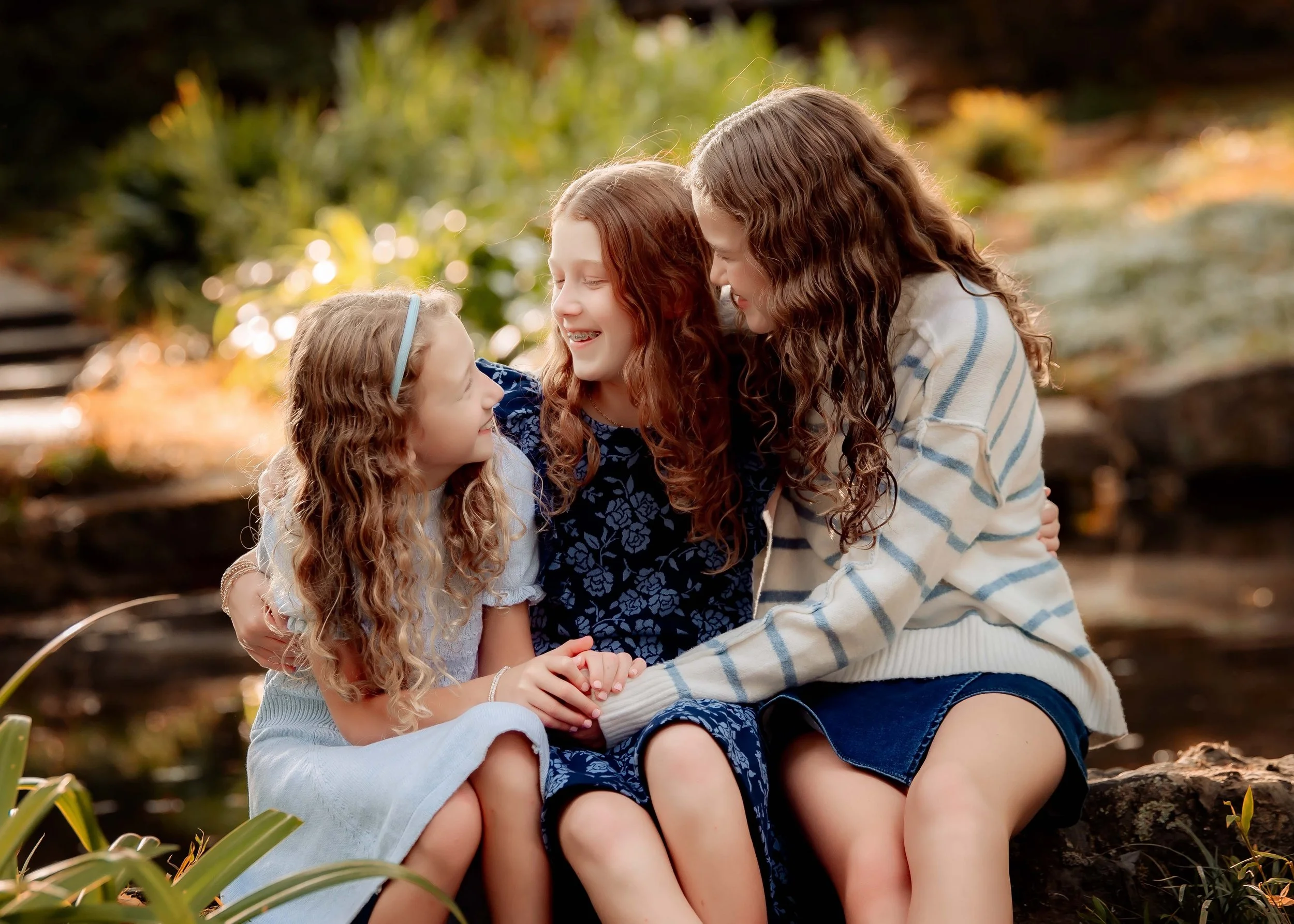 Three girls sitting outdoors by a pond, smiling and holding hands, surrounded by greenery.