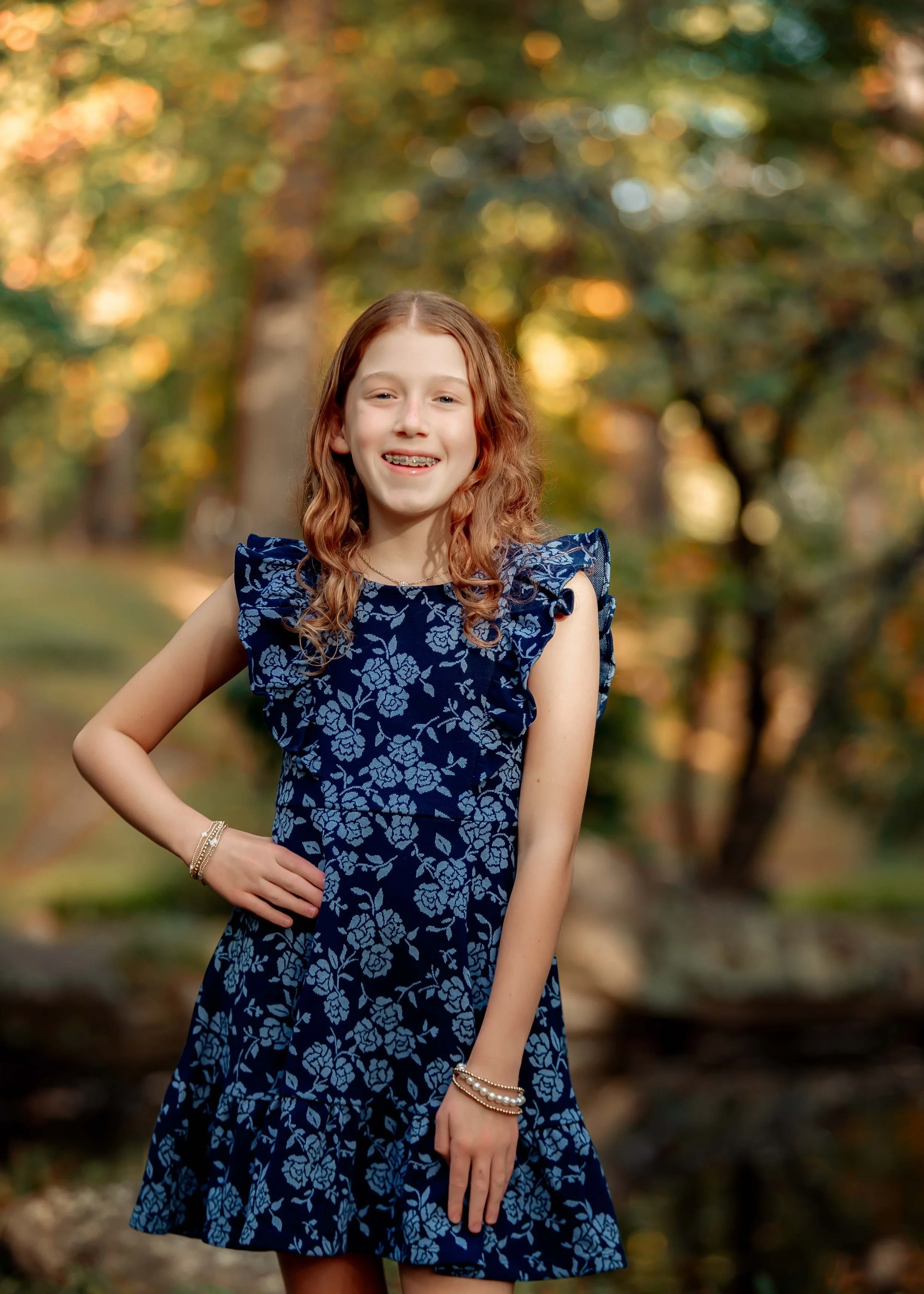 A young girl with long, curly red hair, wearing a blue floral dress with ruffled sleeves, standing outdoors in a park during autumn, smiling at the camera.