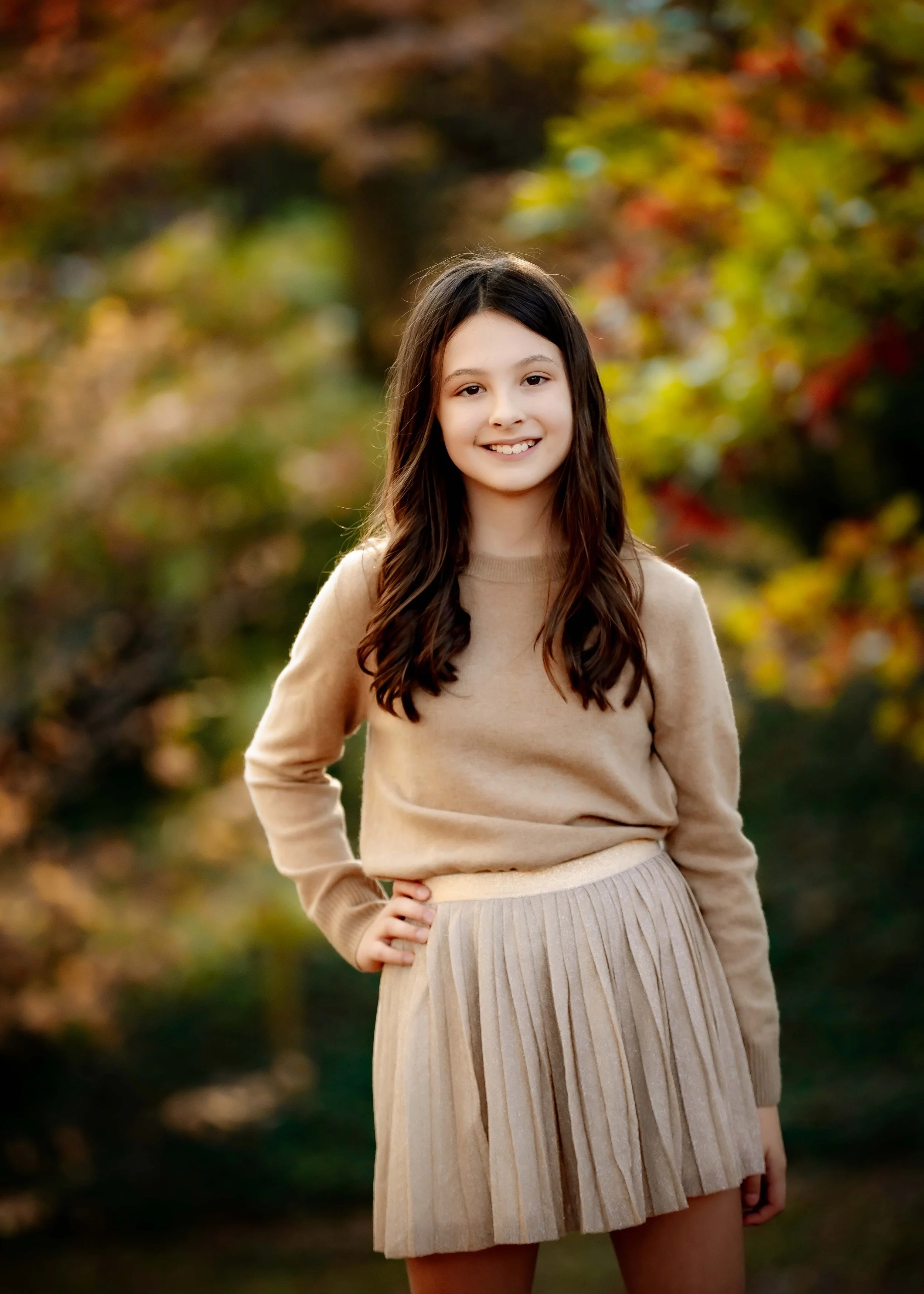 A young girl with long brown hair smiling outdoors during autumn, wearing a beige sweater and pleated skirt.