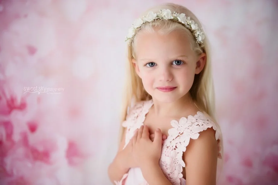 A young girl with long blonde hair wearing a floral headband and a pink lace dress, standing against a soft pink and white background.