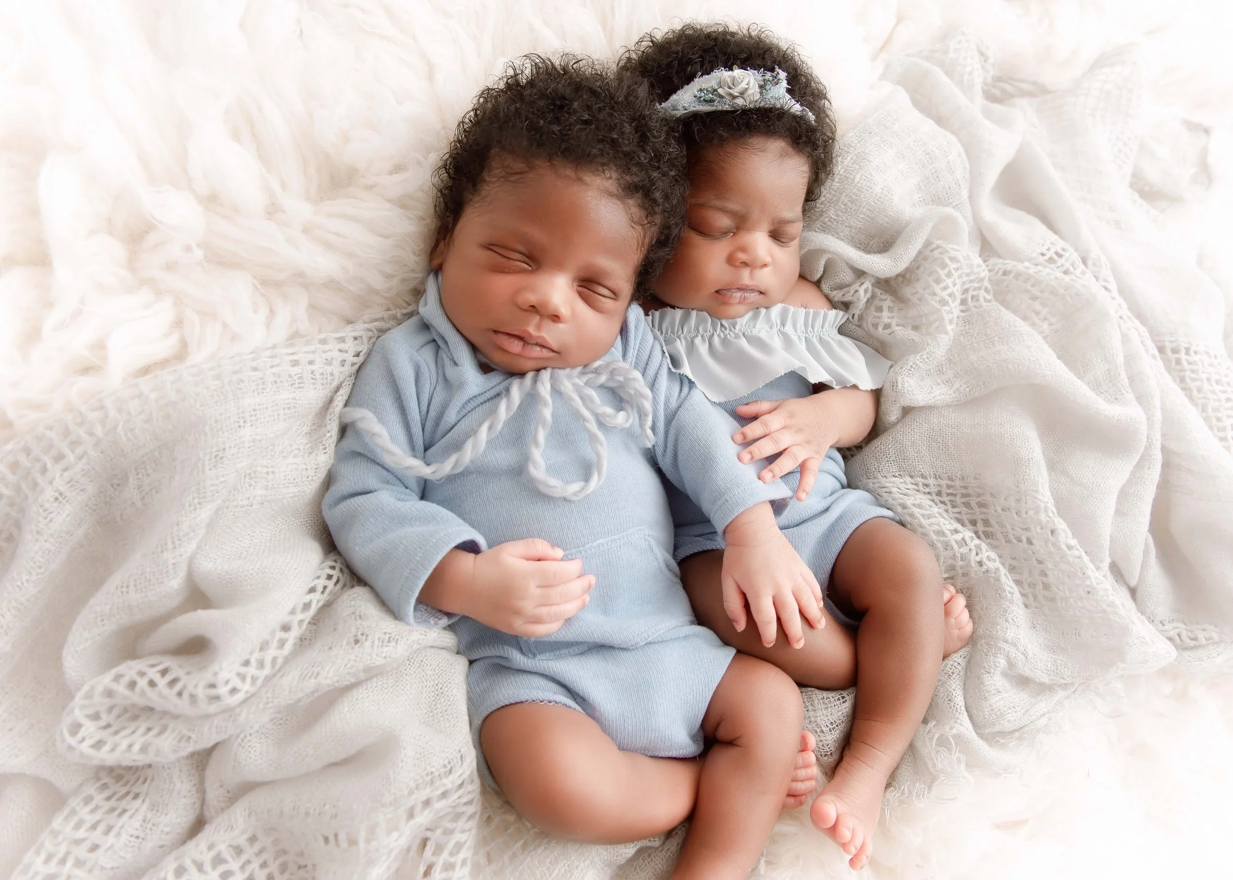Two sleeping infants, a boy and a girl, snuggling on a soft, textured white blanket.