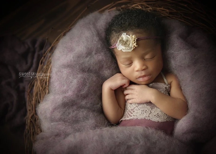 A sleeping baby girl with a headband, lying in a basket filled with soft purple and gray bedding.