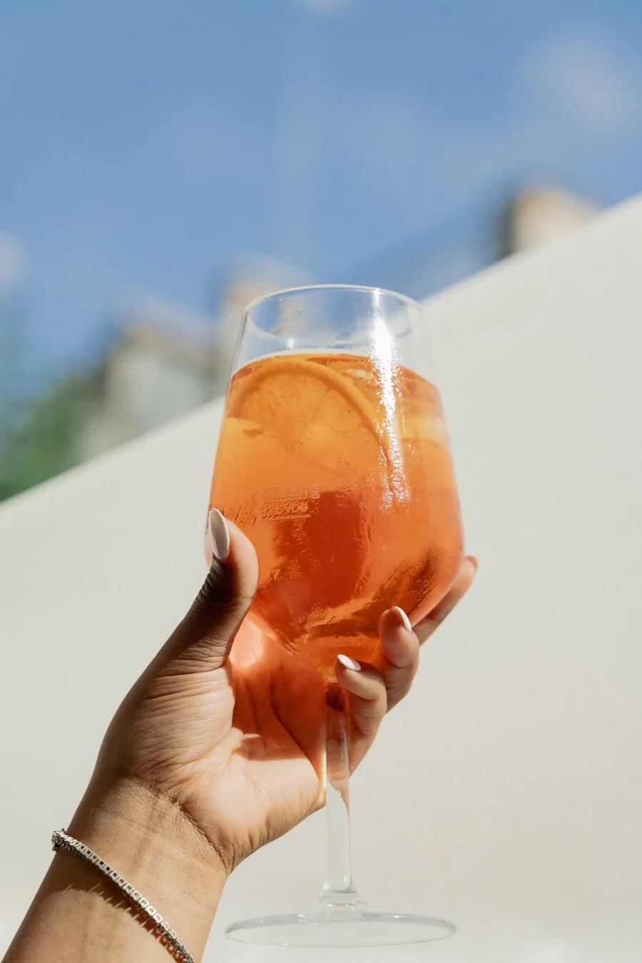 A person holding a wine glass filled with an orange-colored drink and slices of oranges inside, with a blue sky background.