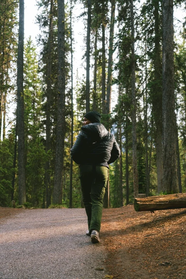 Person walking on a trail in a forest, wearing a black jacket, green pants, and a black beanie, surrounded by tall trees.