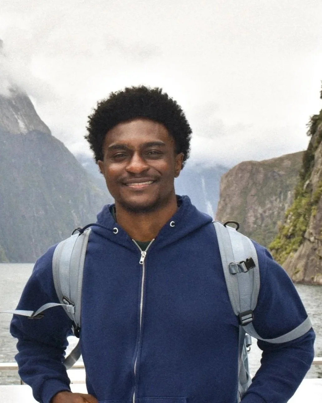 Man smiling outdoors in a scenic mountainous area with water and waterfalls in the background