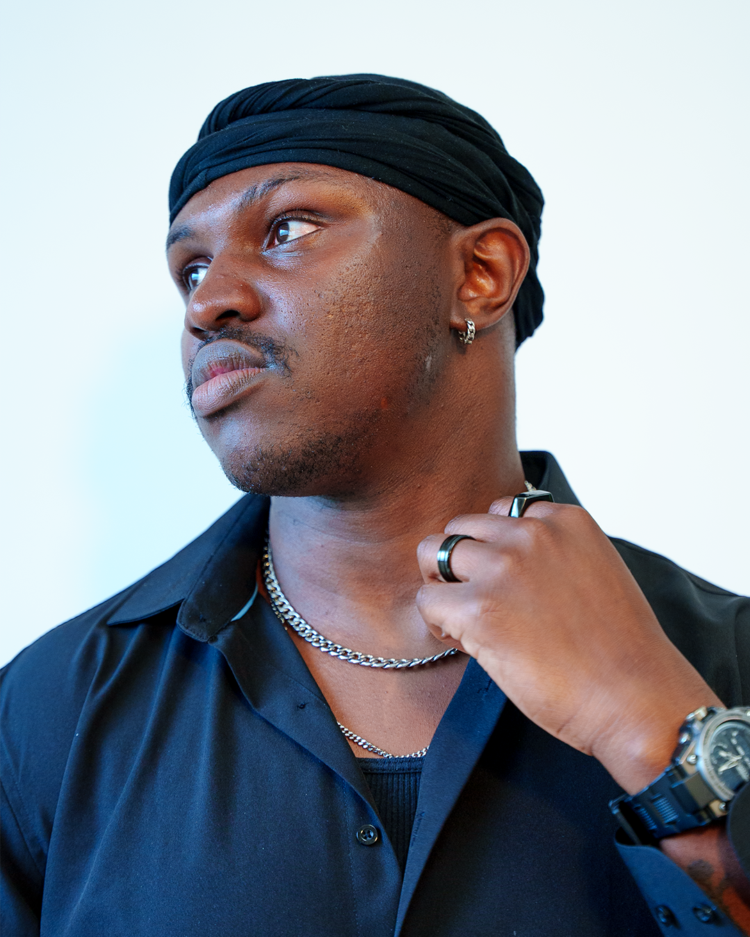 Portrait of a man wearing a black headscarf, black shirt, silver chains, earrings, ring, and watch, looking to the side with his hand on his neck.