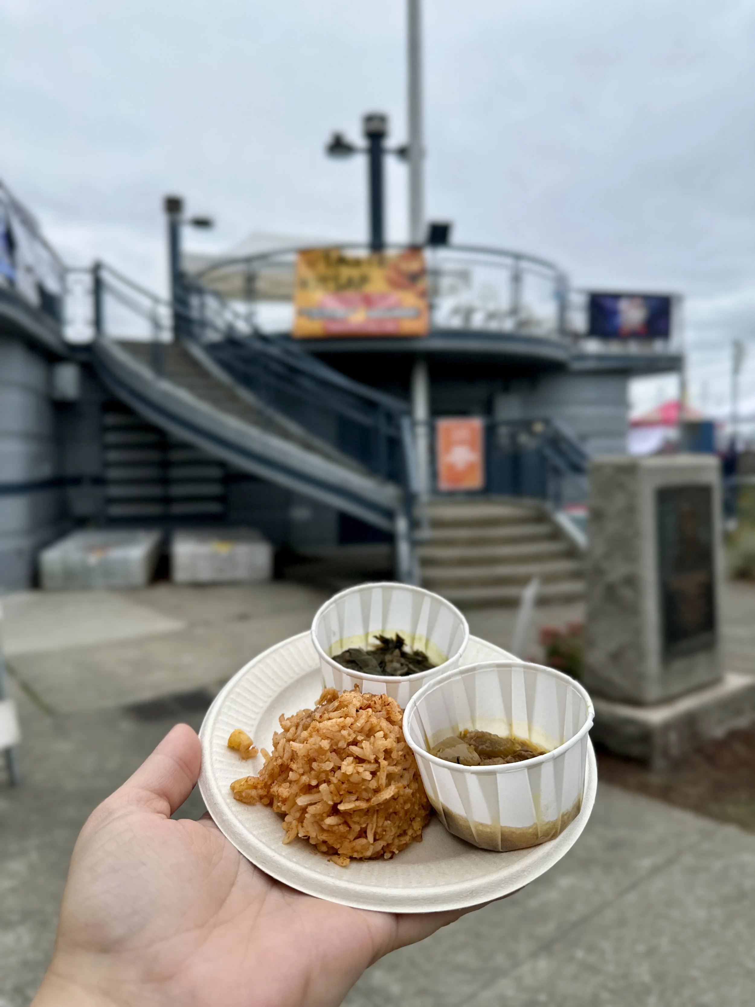 A person's hand holding a paper plate with fried rice, and three cups of food, with an outdoor setting featuring a staircase, railings, and billboards in the background.
