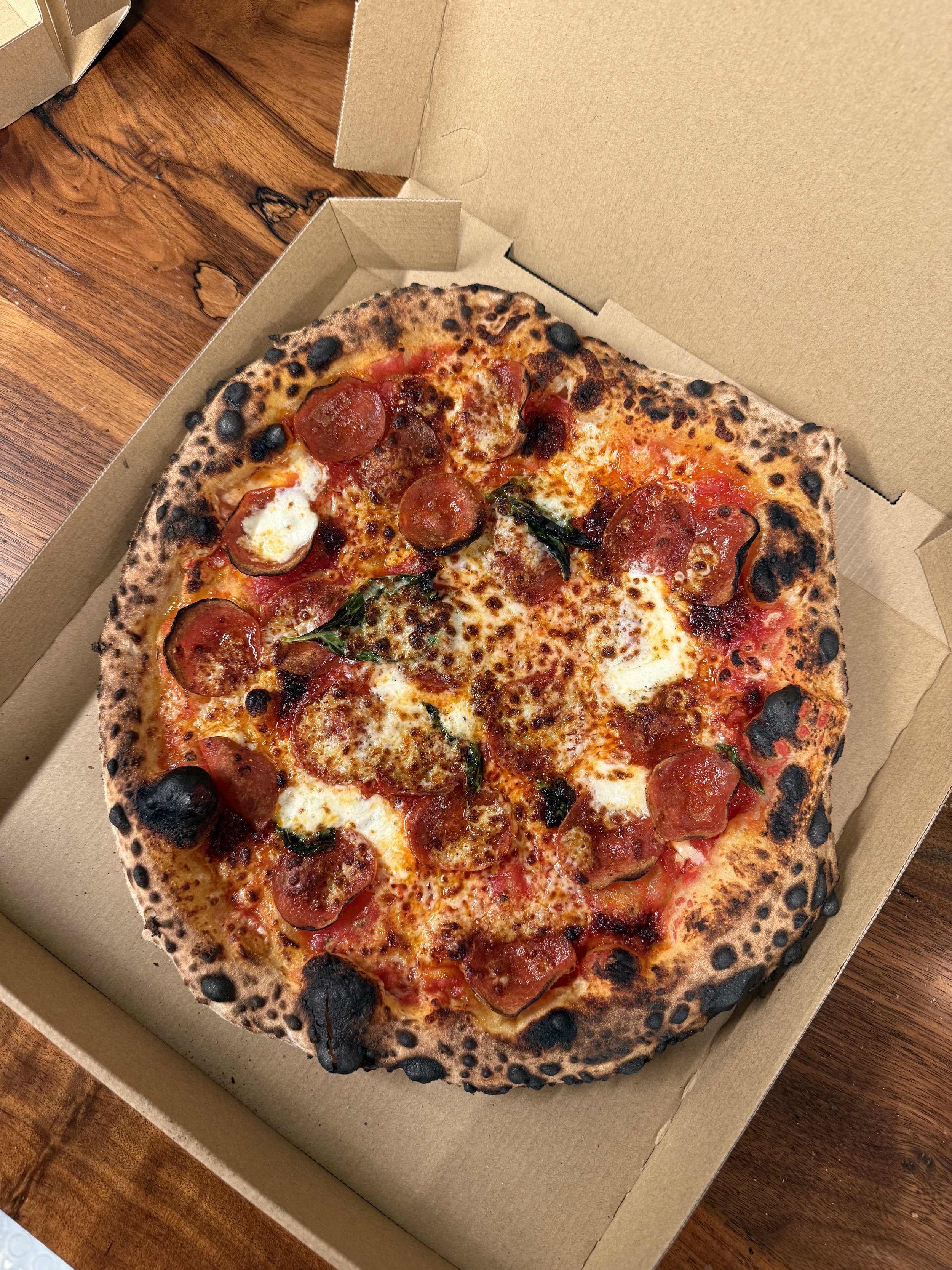 A large pepperoni pizza with some charred spots on the crust inside an open cardboard pizza box on a wooden table.